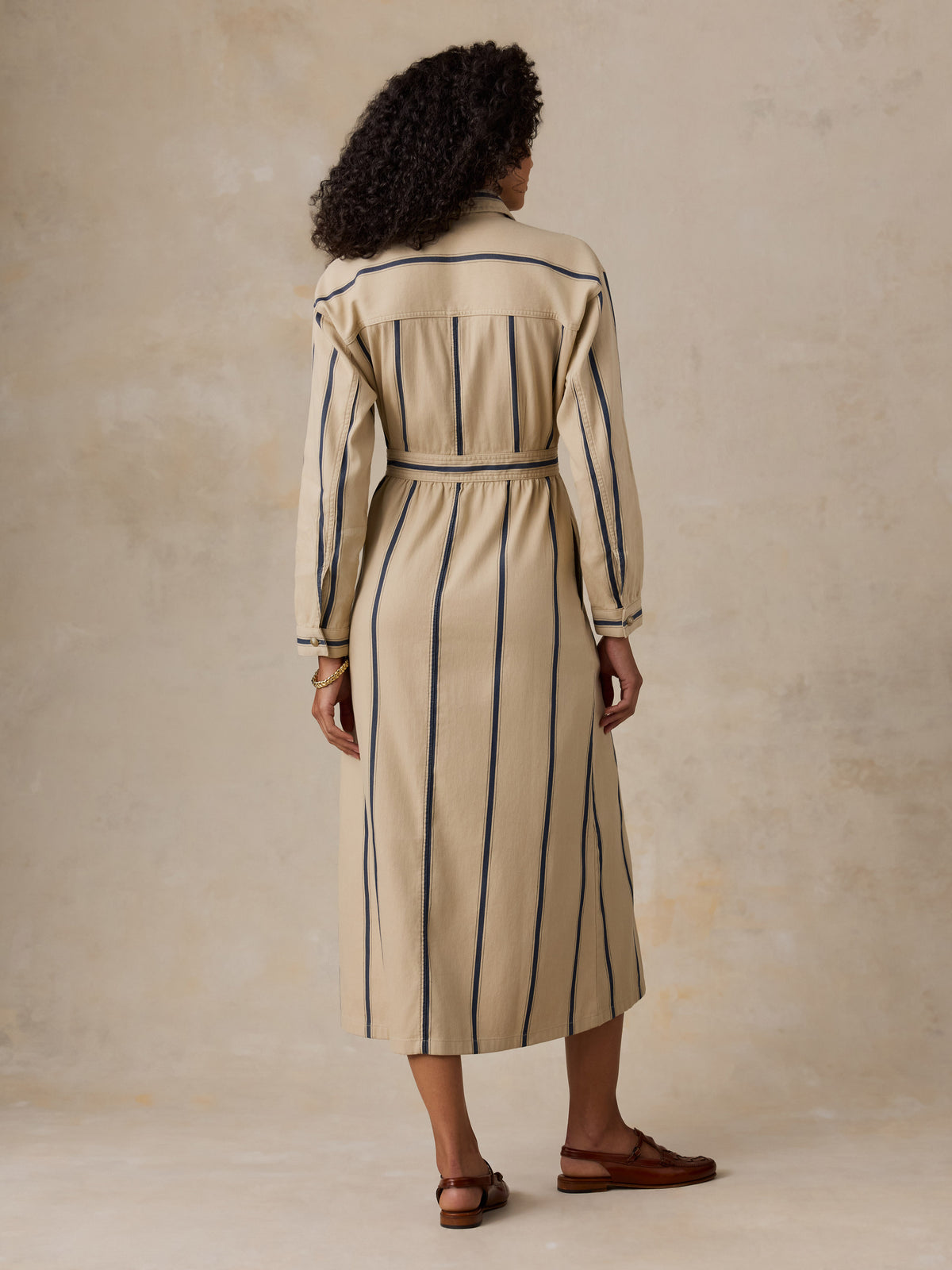 A woman viewed from behind wearing a beige midi shirt dress with vertical navy blue stripes and a matching waist tie, paired with brown leather sandals, set against a textured beige studio backdrop., image 3 of 4
