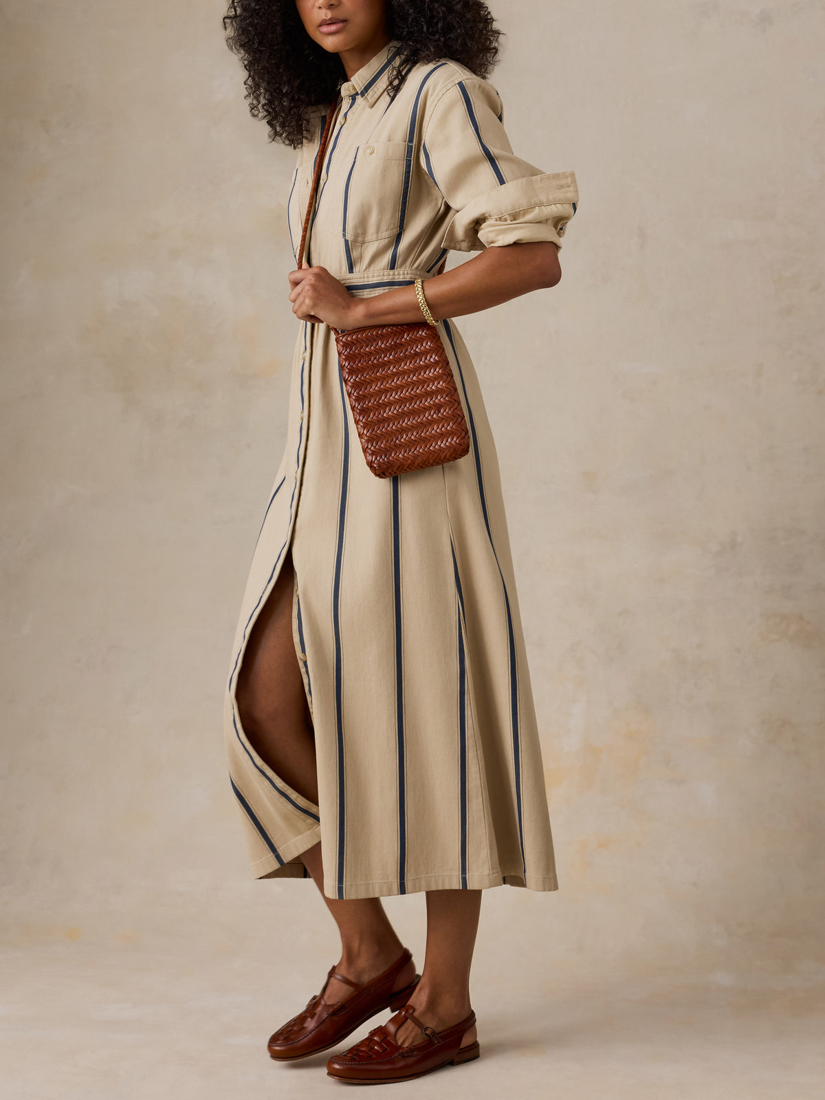 A woman models a beige midi shirt dress with vertical navy blue stripes, featuring a high side slit, puffed short sleeves, and a button-down collar. She is holding a brown woven crossbody bag and wearing brown leather loafers, set against a textured beige studio backdrop., image 4 of 4