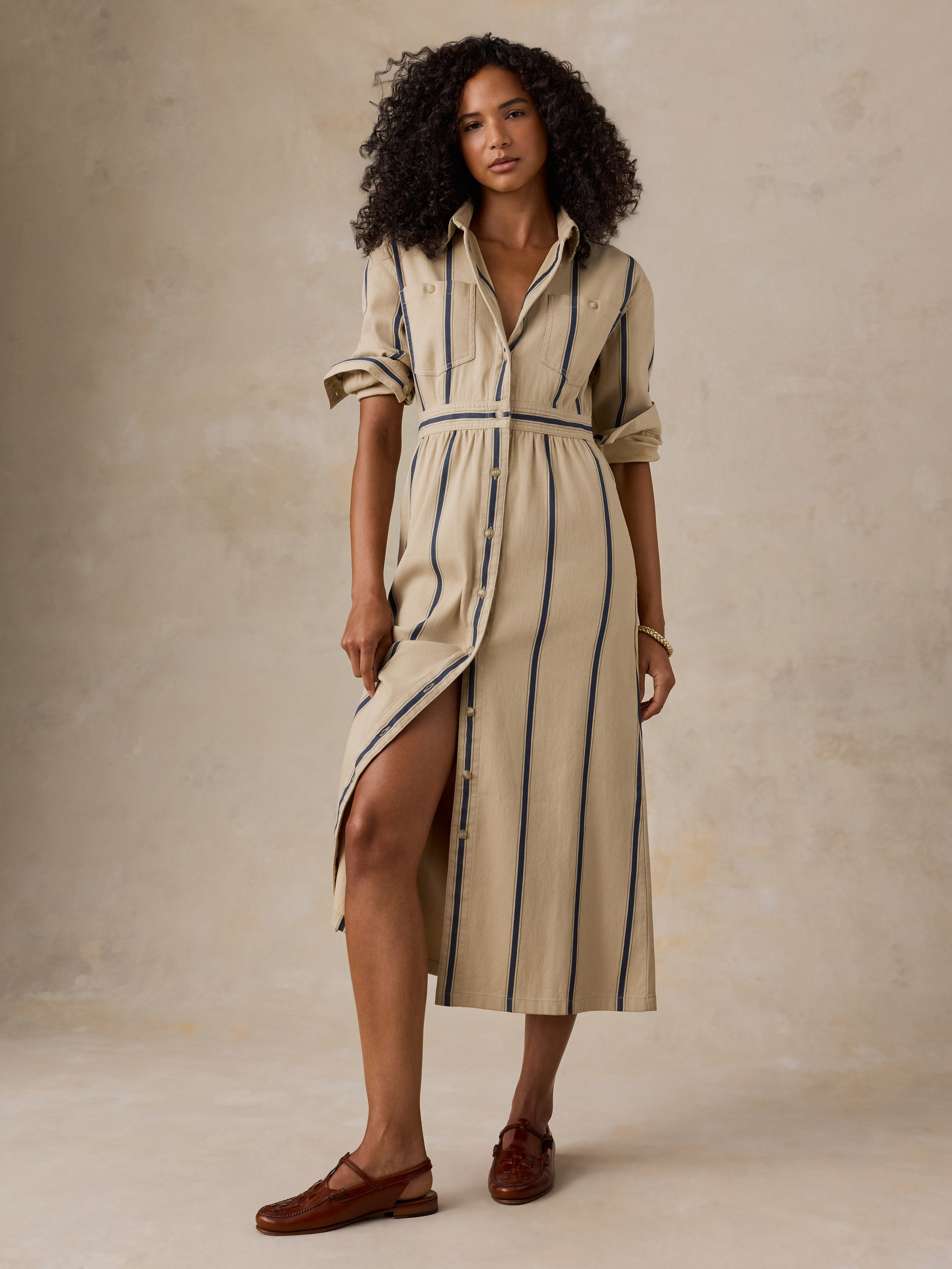 A woman models a beige midi shirt dress with vertical navy stripes, rolled sleeves, and a button-down front, paired with brown leather loafers against a textured beige studio backdrop.