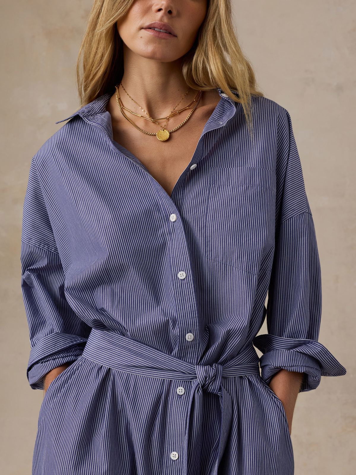 A woman wearing a blue and white vertically striped button-down shirt dress with a tie waist belt, styled with layered gold necklaces against a textured beige background., image 2 of 4