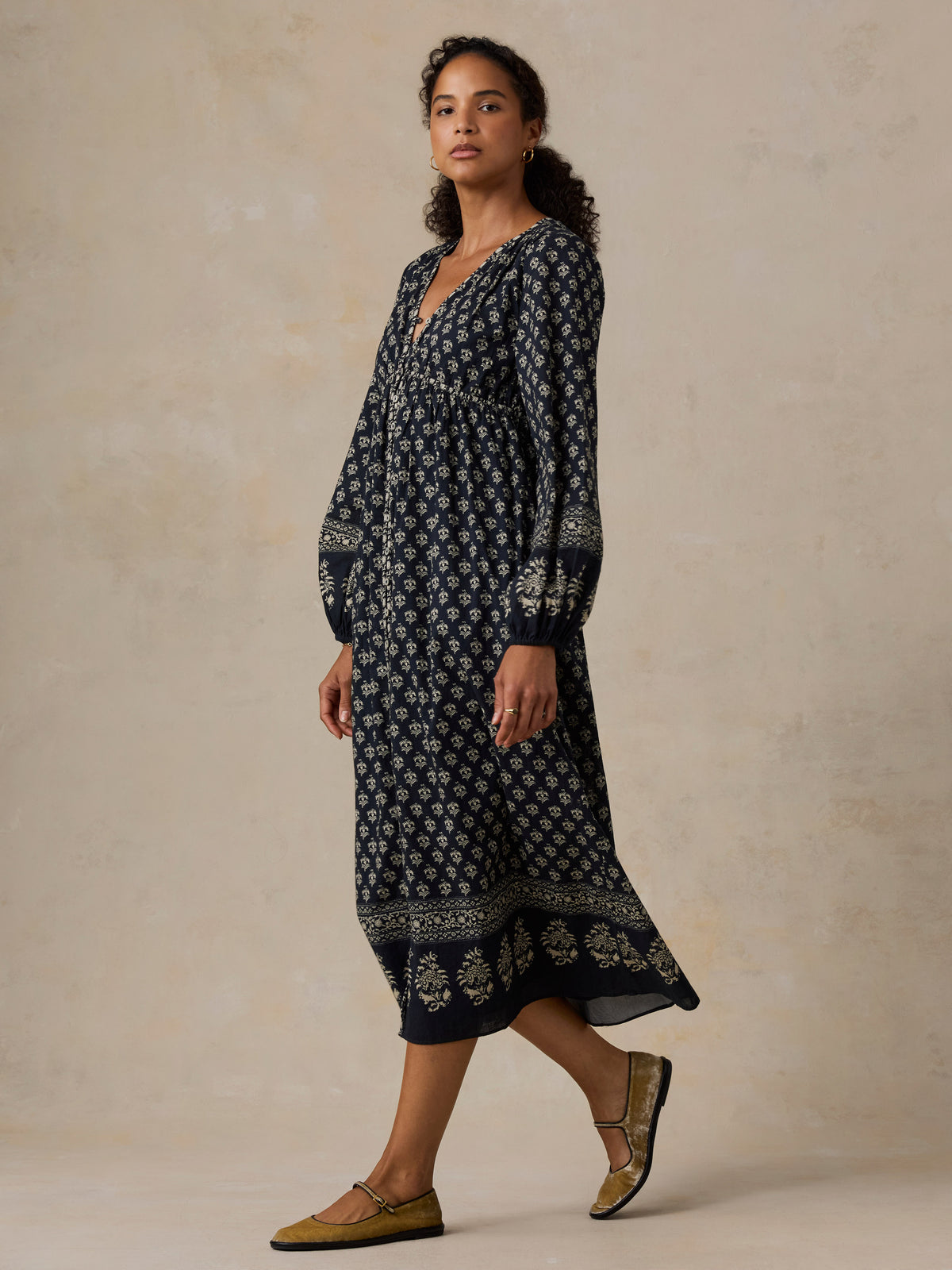 A woman modeling a long-sleeve navy midi dress with a white floral block print and a decorative border, paired with olive green flat sandals, set against a textured beige studio backdrop., image 1 of 4