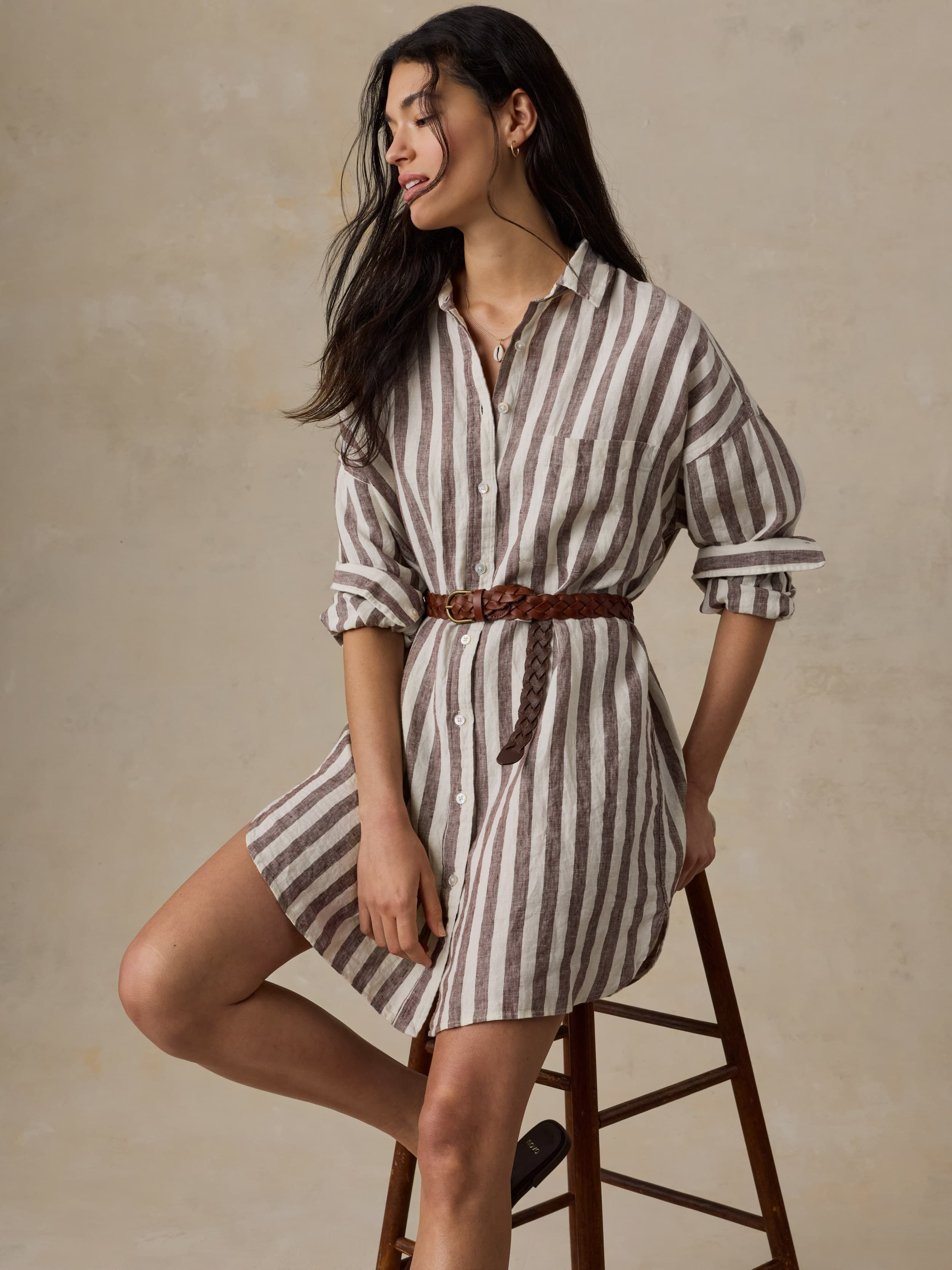Woman wearing a striped shirt dress sitting on a wooden stool against a beige background