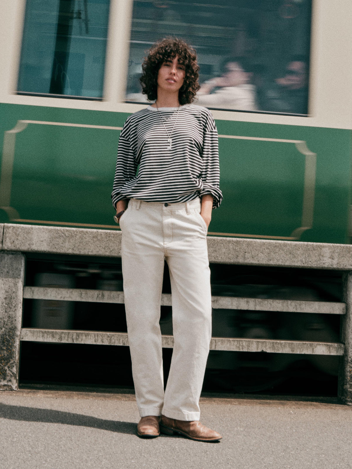 A woman stands outdoors in front of a green and cream train, wearing a black and white striped long-sleeve top, off-white trousers, and brown leather shoes., image 2 of 9
