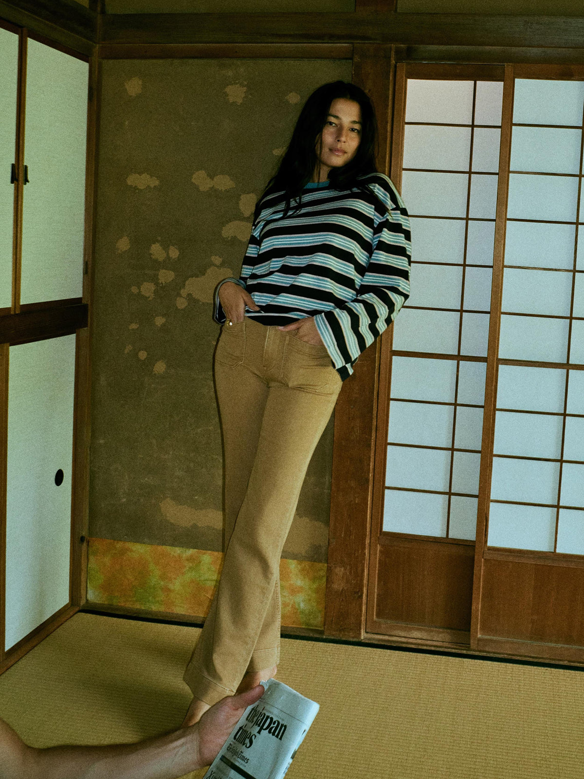 A woman stands in a traditional Japanese room wearing a blue and white striped long-sleeve top and tan pants. A hand in the bottom-left corner holds a newspaper., image 3 of 8