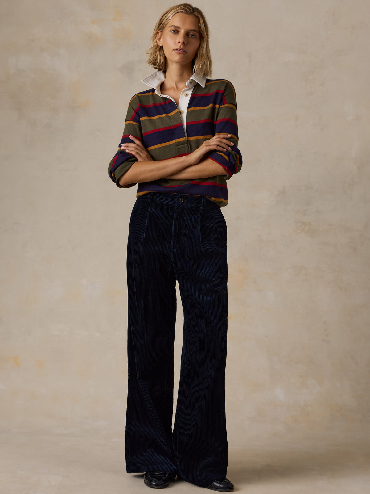A woman wearing a long-sleeve rugby-style shirt with stripes in olive green, navy blue, red, and mustard yellow, image 5 of 7