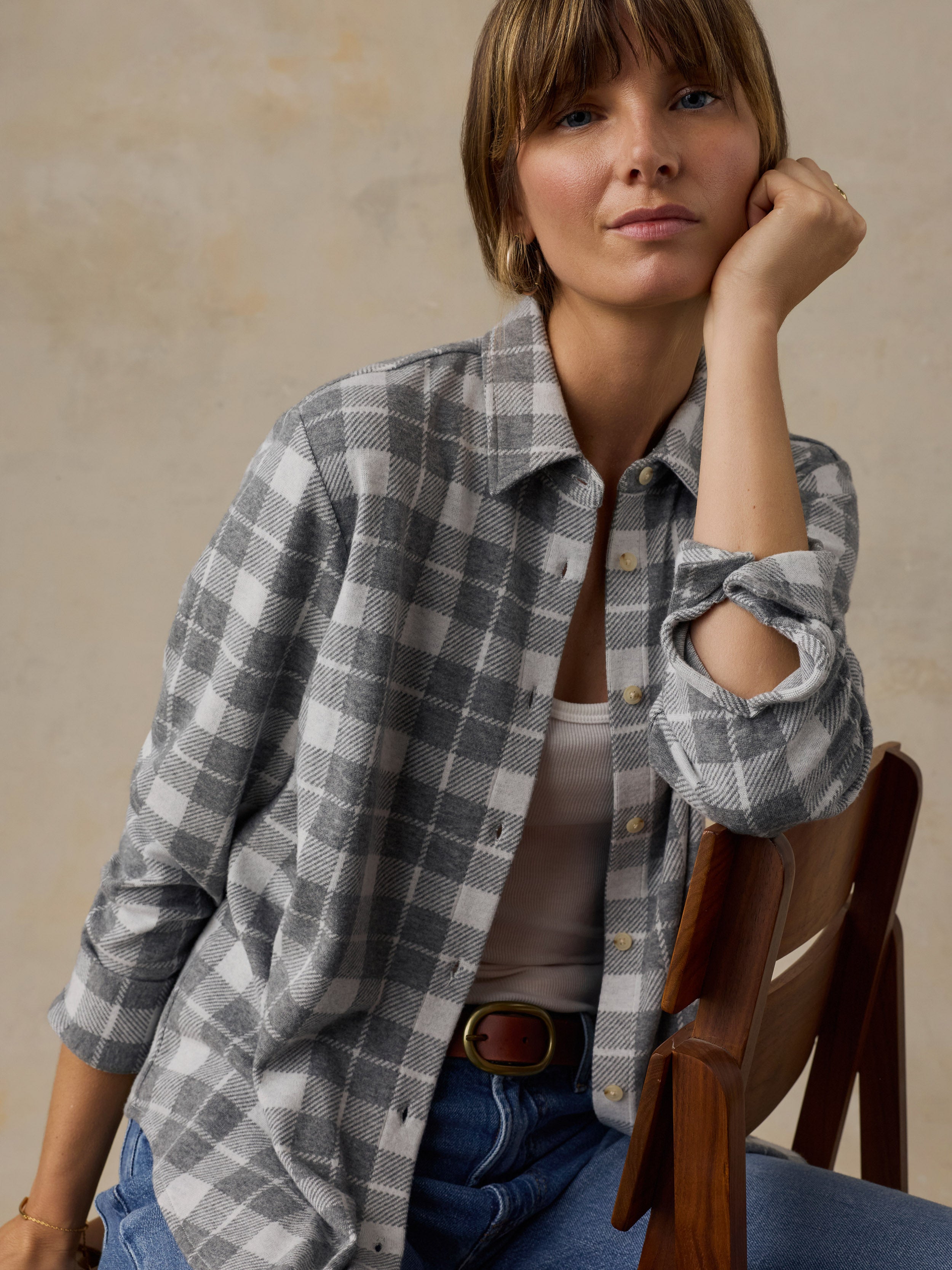 Woman sitting on a wooden chair wearing a grey plaid button down shirt and blue jeans against a beige background
