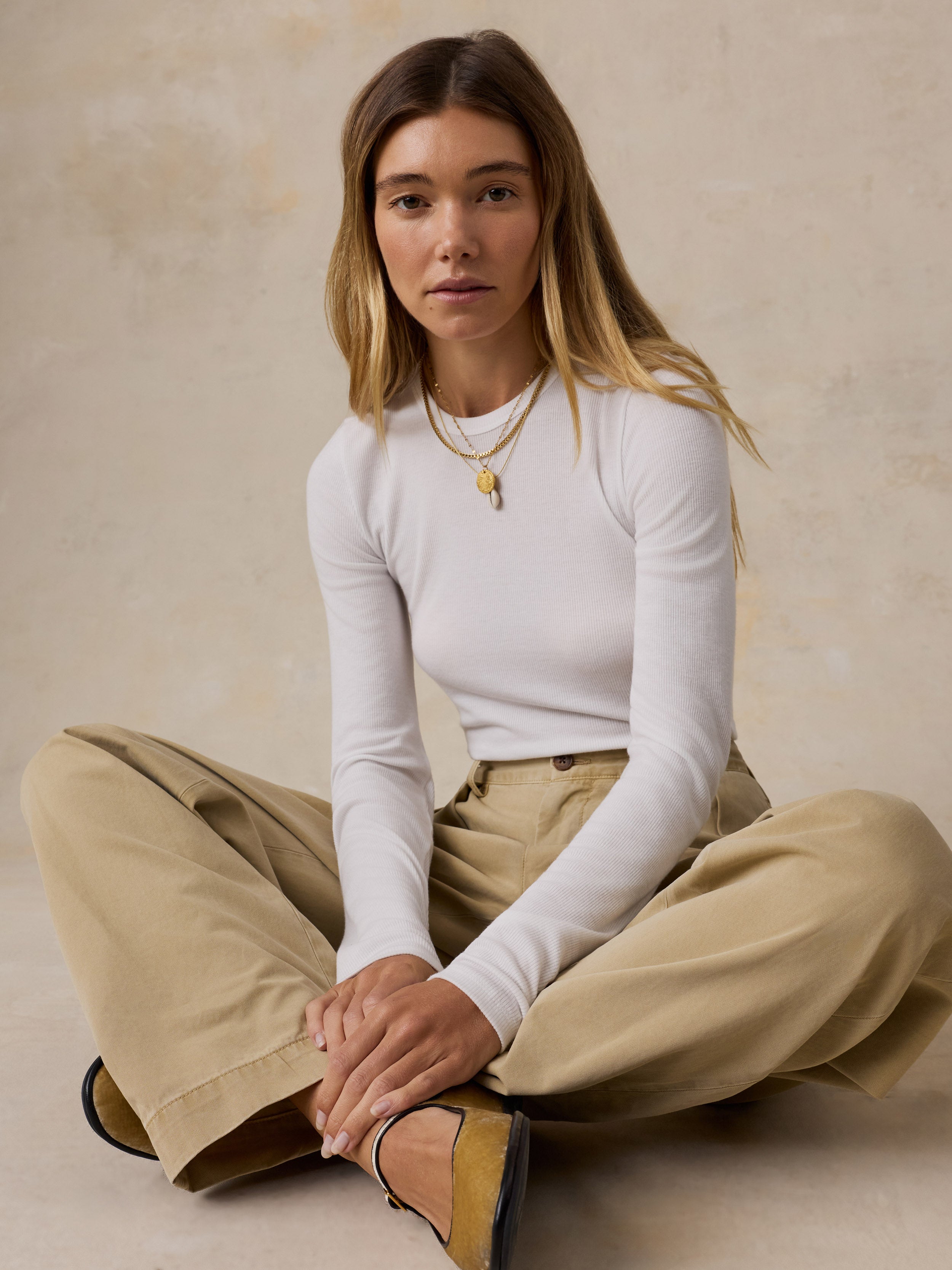 A woman sits cross-legged on a textured beige studio floor wearing a fitted white long-sleeve top, loose beige wide-leg pants, and tan flats, with layered gold necklaces.