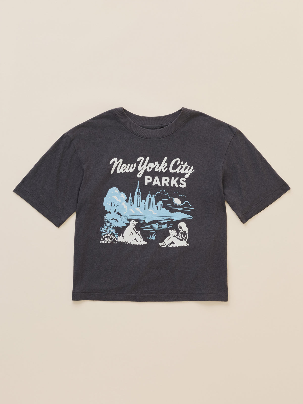 A dark charcoal gray short-sleeve t-shirt with a 'New York City Parks' graphic on the front, featuring a light blue and white illustration of a park scene with a skyline, laid flat against a warm beige background., image 6 of 6