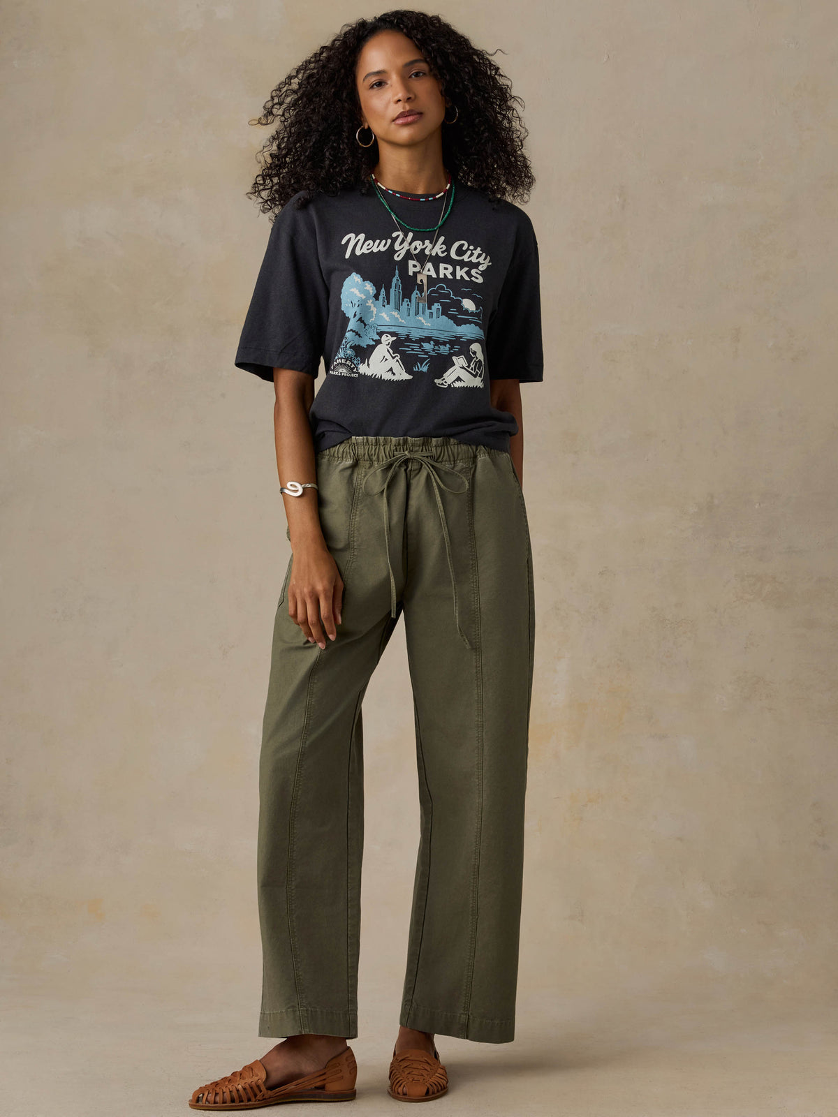 Woman wearing a black 'New York City Parks' t-shirt and green pants on a beige background, image 5 of 6