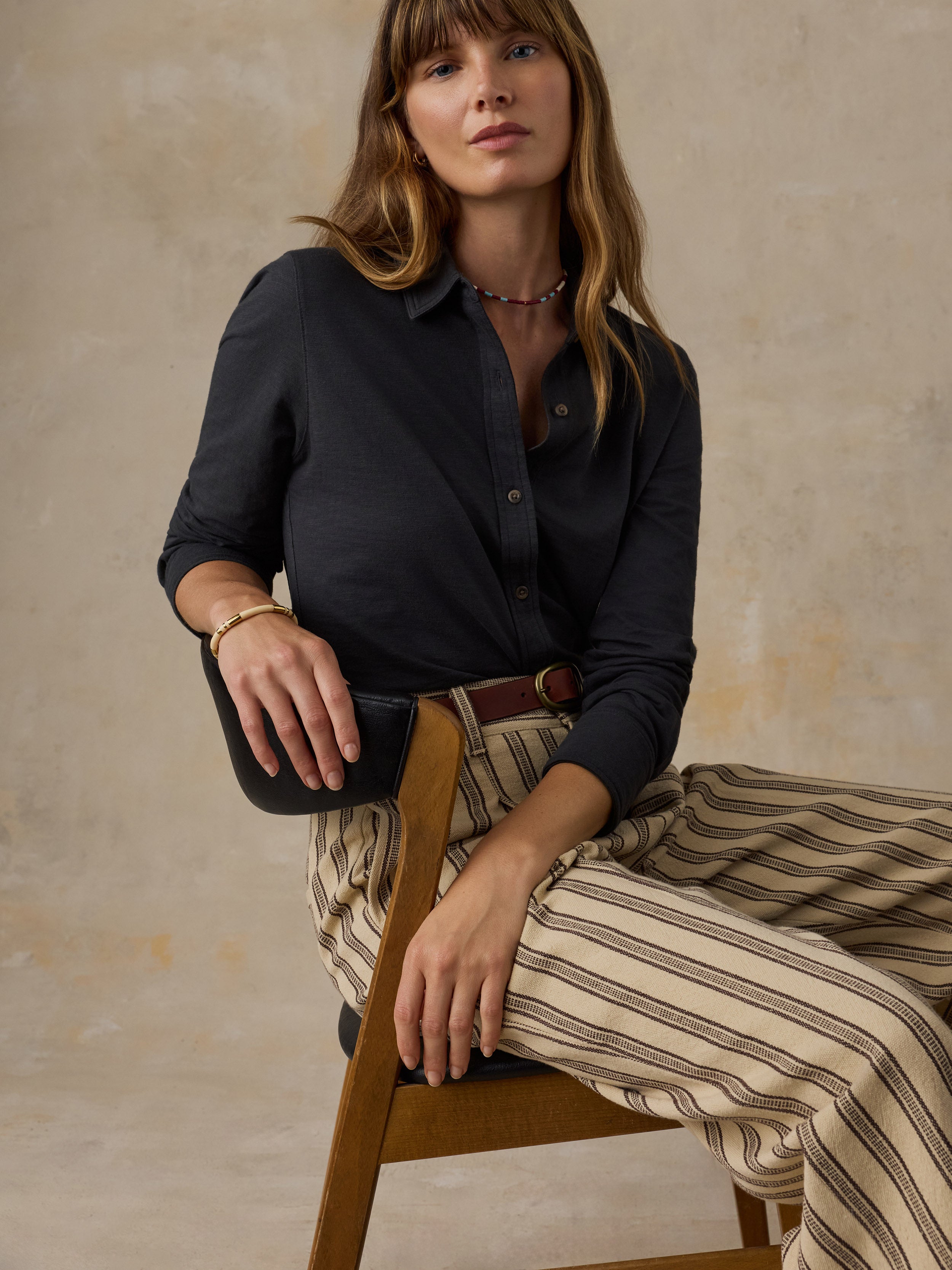 Woman sitting in a wooden chair wearing a dark charcoal button up shirt and striped pants against a beige background