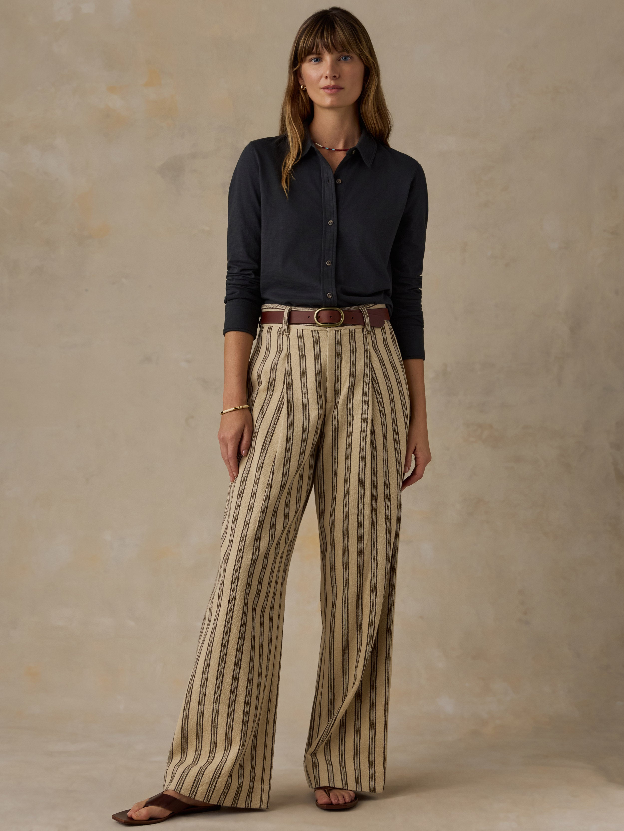 Woman wearing a dark charcoal button up shirt and striped pants against a beige background