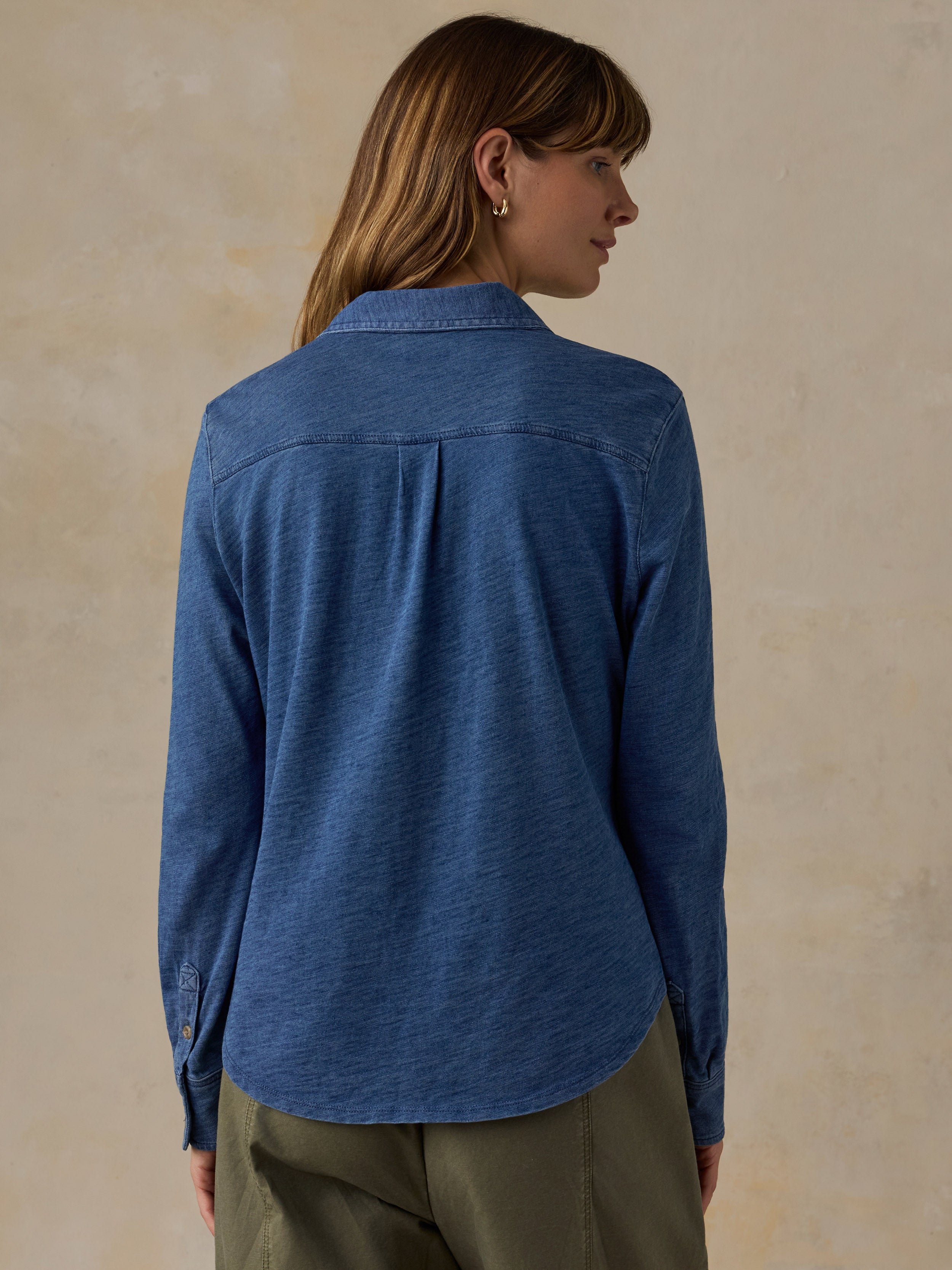 Woman wearing a blue button down shirt and olive green pants against a beige background