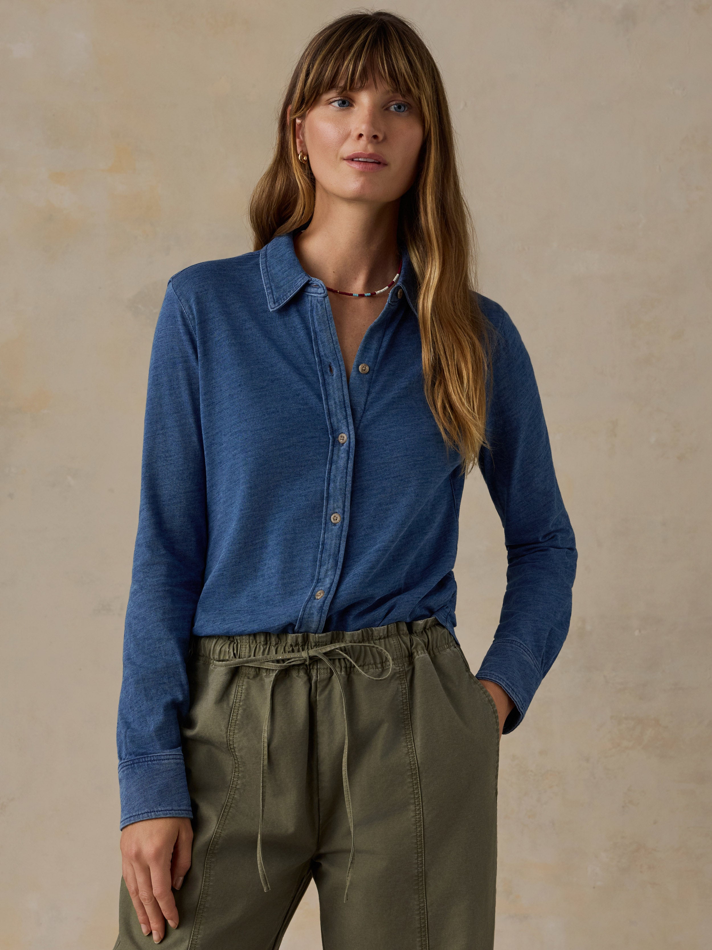 Woman wearing a blue button down shirt and olive green pants against a beige background
