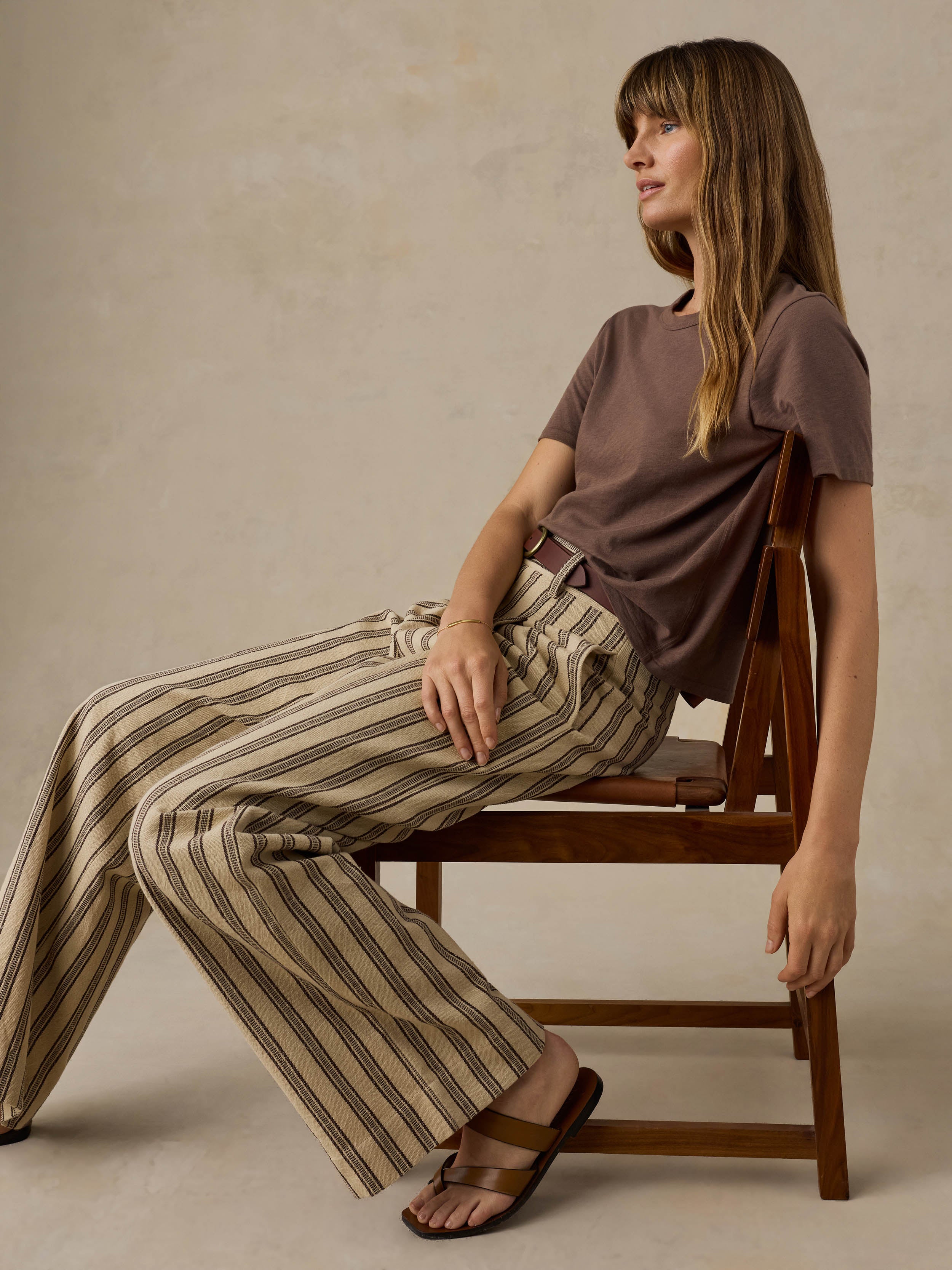 Woman sitting on a wooden chair wearing a brown t-shirt and striped pants against a beige background
