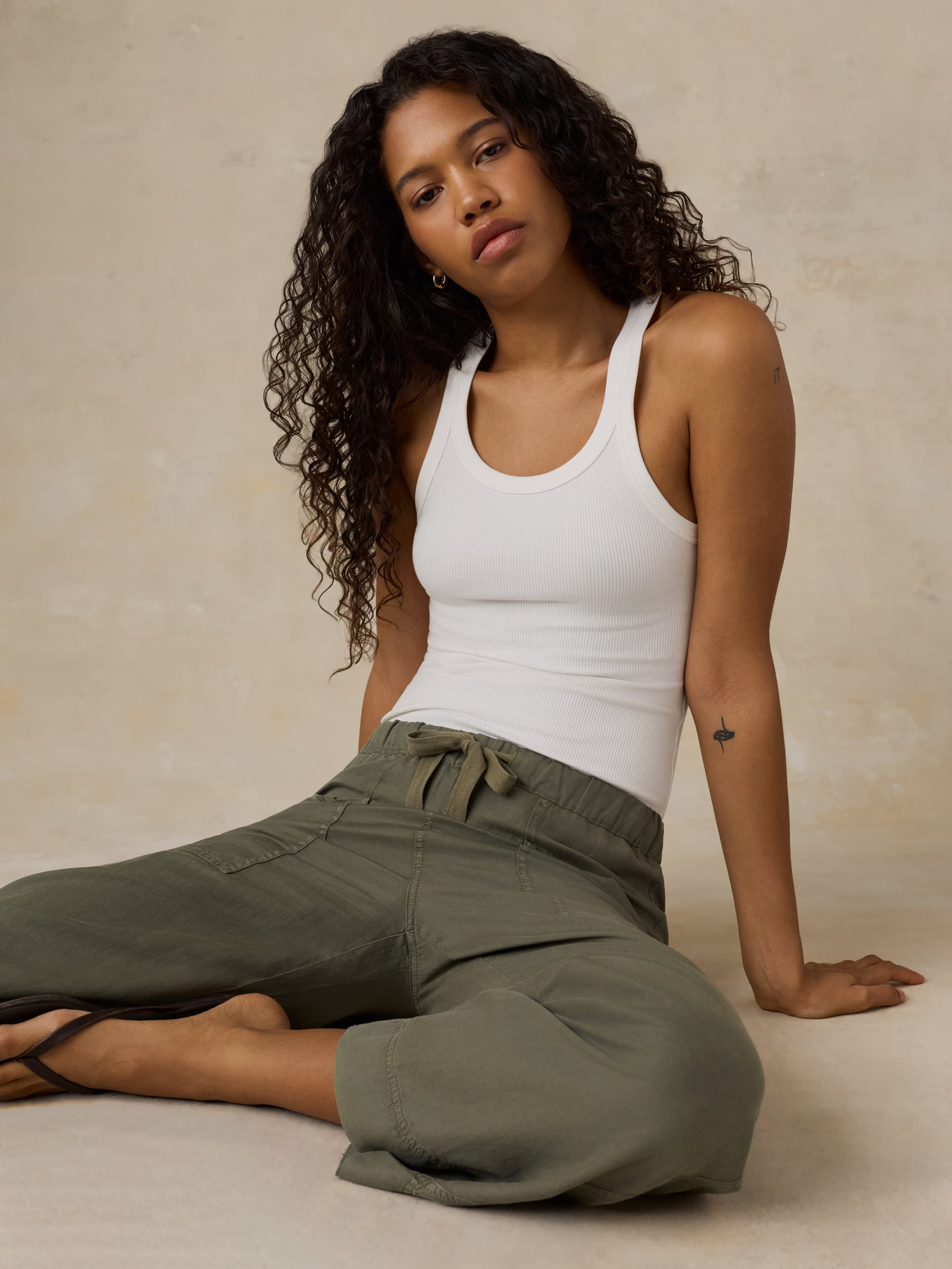 Woman wearing a white tank top and green pants sitting on a beige background