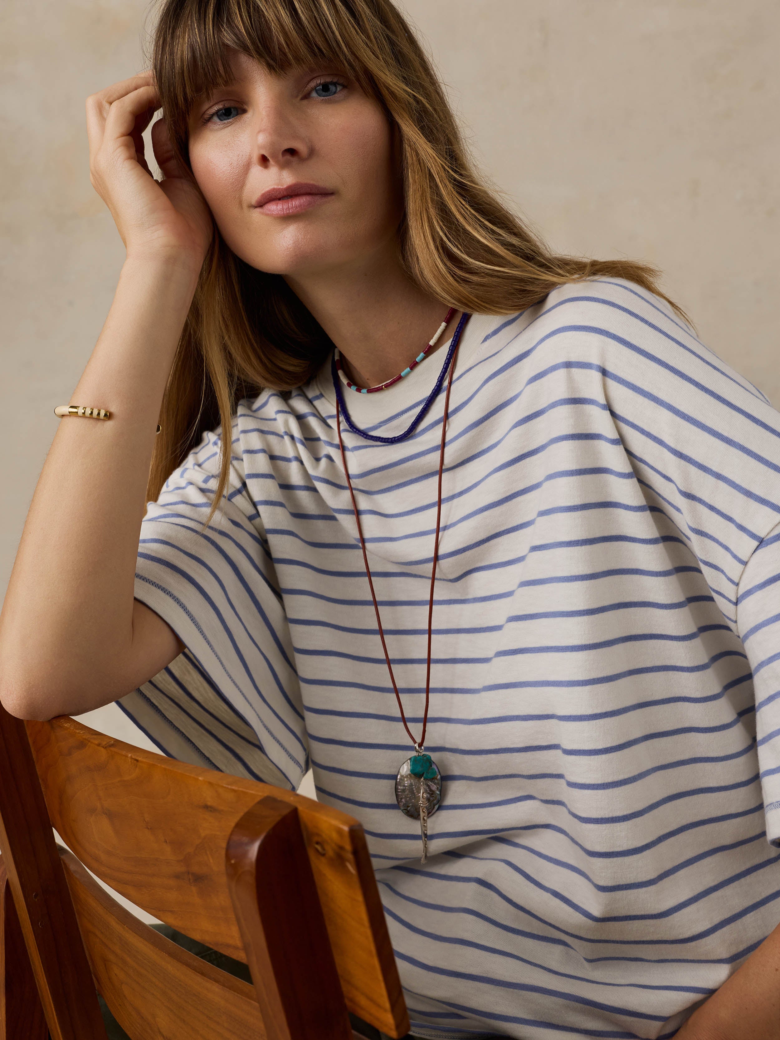 Woman sitting on a wooden chair wearing blue and white striped t-shirt against a beige background