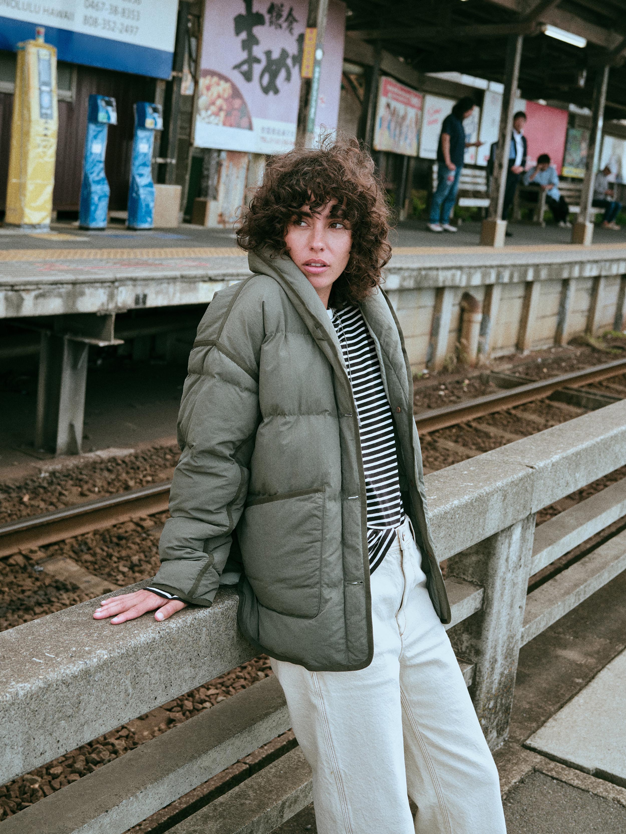 A woman wearing an olive green puffer jacket over a black and white striped shirt and white pants.