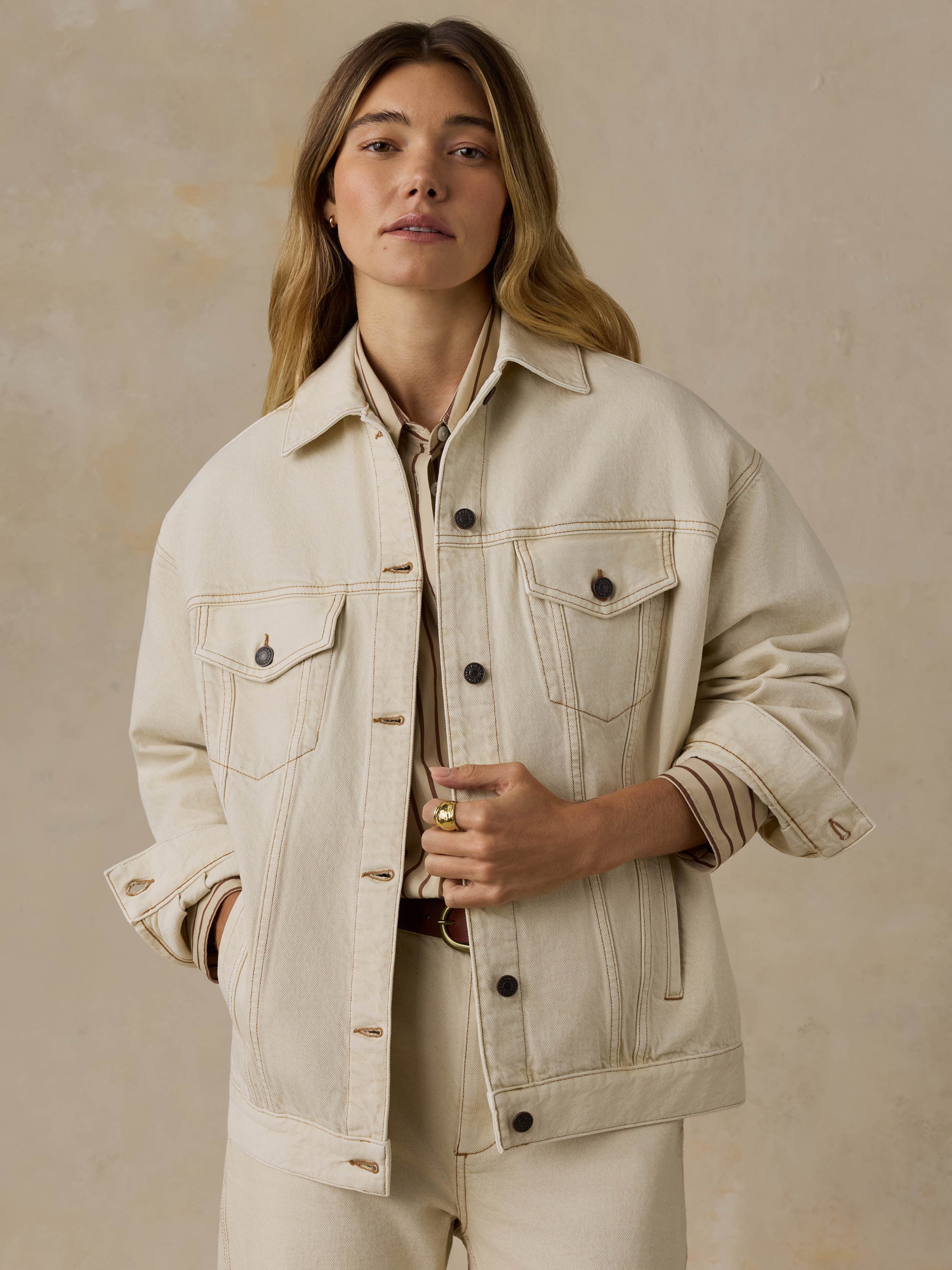 Woman wearing an off white denim jacket and pants against a beige background