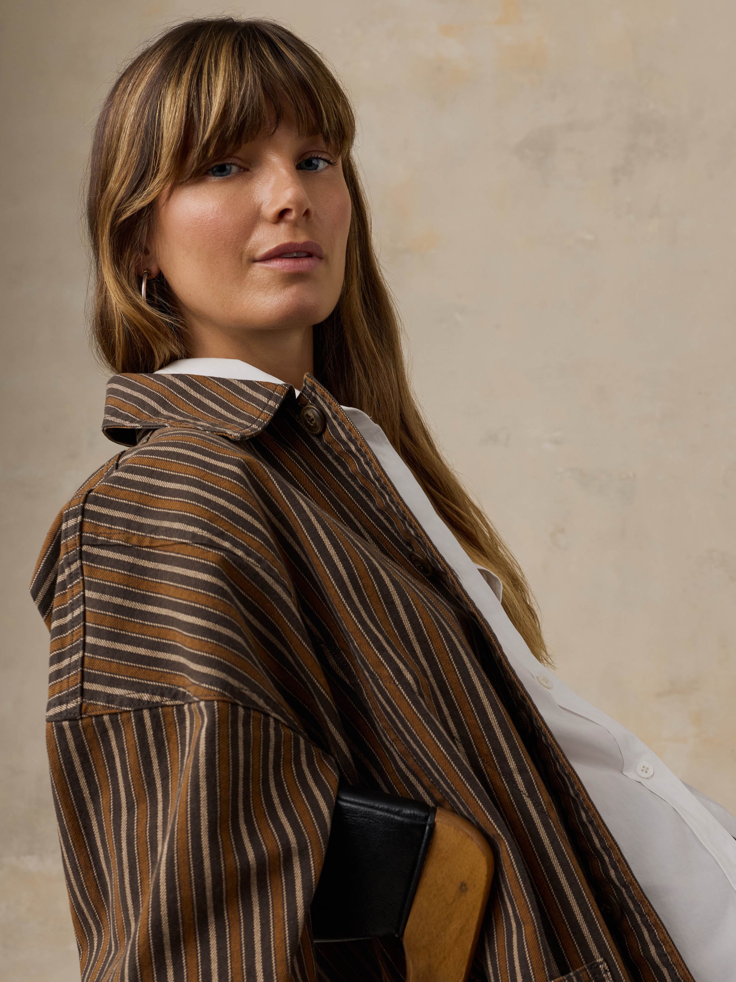 Woman sitting on a wooden chair wearing a brown striped chore jacket against a beige background