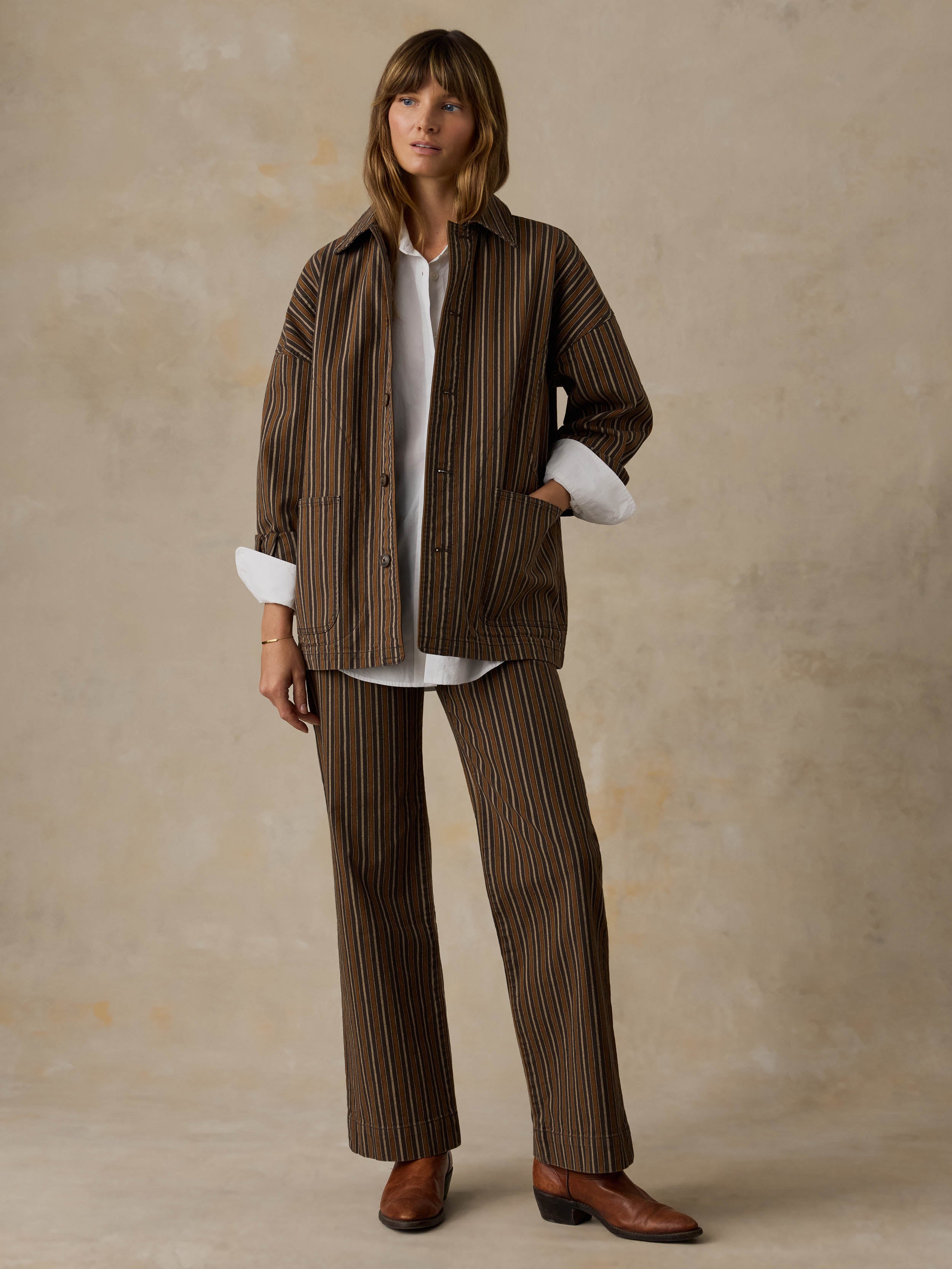 Woman wearing a white button down shirt, brown striped chore jacket, and matching striped pants against a beige background