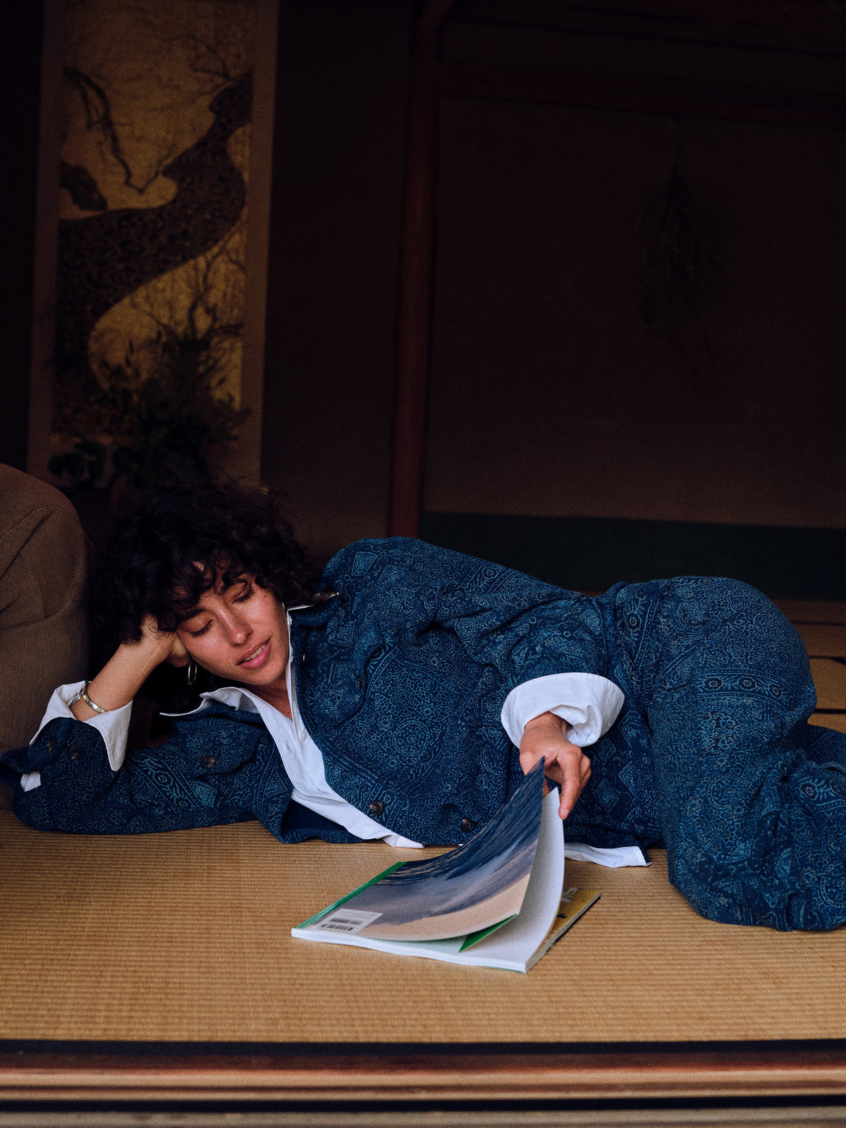 A woman reclines on a tatami mat floor wearing a dark blue patterned kimono jacket over a white collared shirt, holding an open magazine in a dimly lit interior setting., image 7 of 7