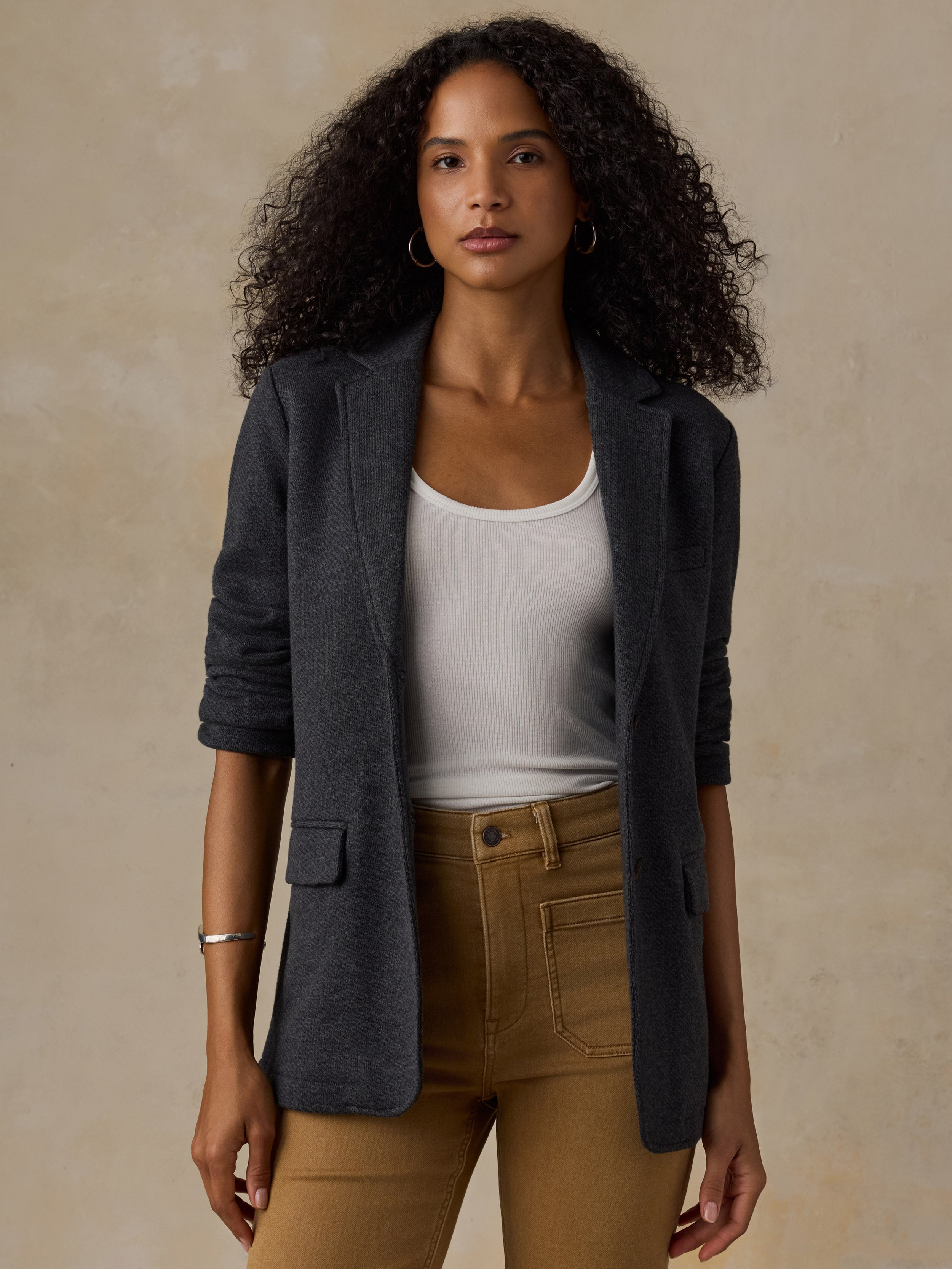 Woman wearing a black blazer over a white tank and brown pants against a beige background