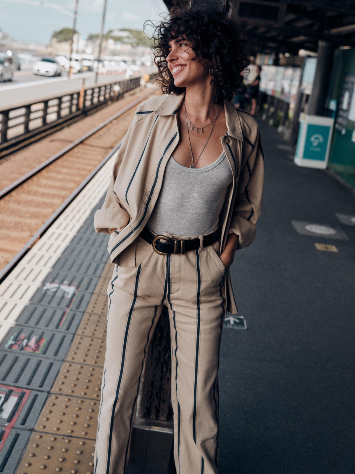 A woman with curly dark hair stands on a train station platform wearing a beige tailored suit with thin dark stripes, a light gray tank top, and a black belt., image 7 of 7