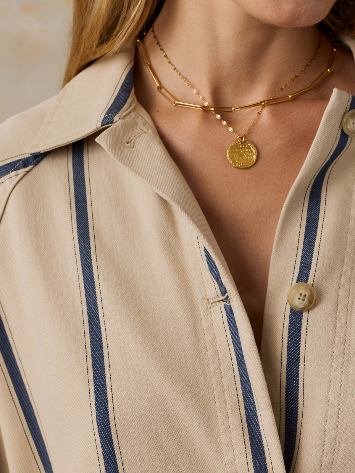 Close-up of a model wearing layered gold necklaces, including a chain with a round coin pendant, over a beige and navy striped button-up shirt., image 4 of 7
