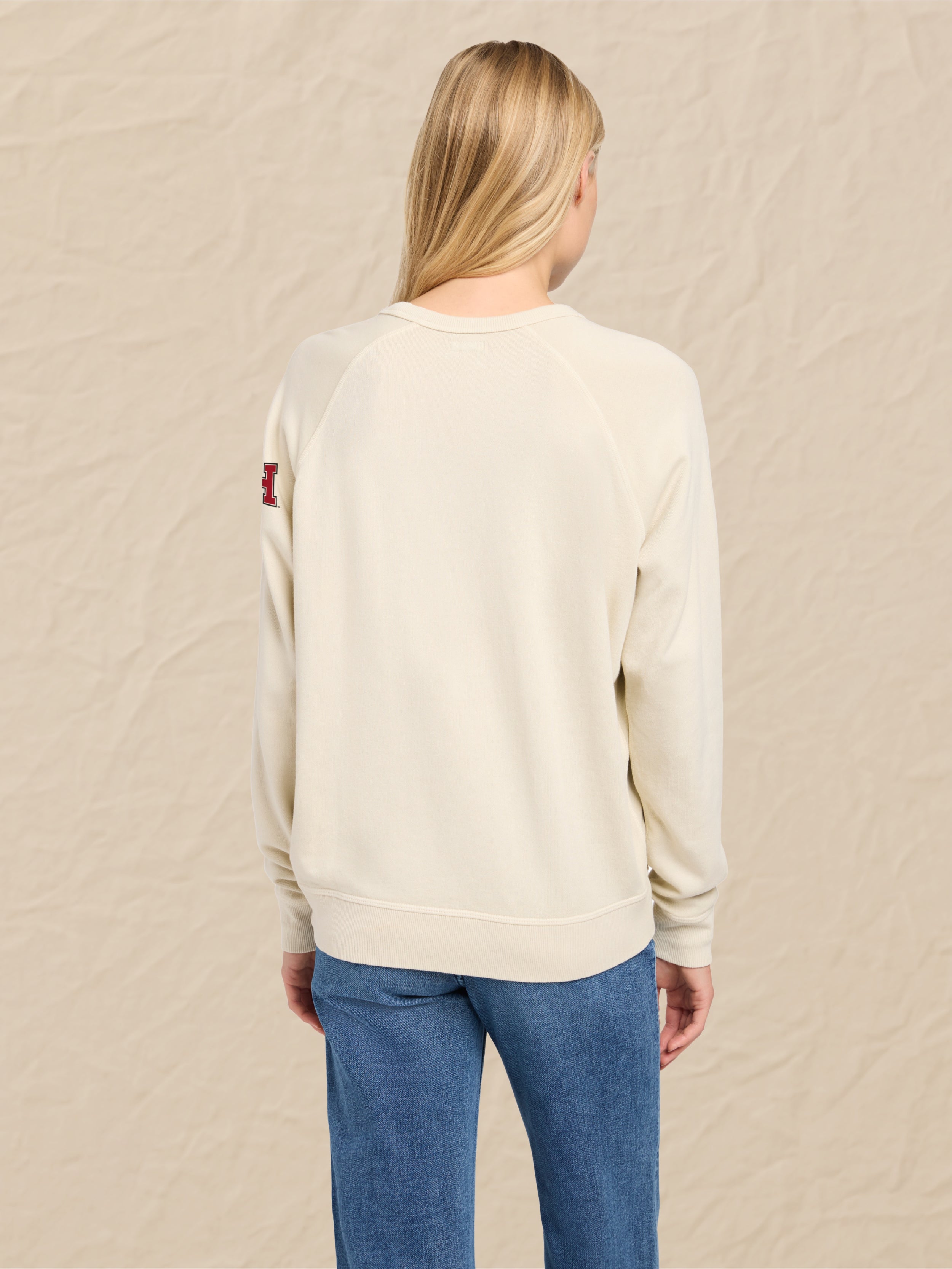 Woman wearing an ivory sweatshirt with Harvard graphic on the left shoulder against a beige background
