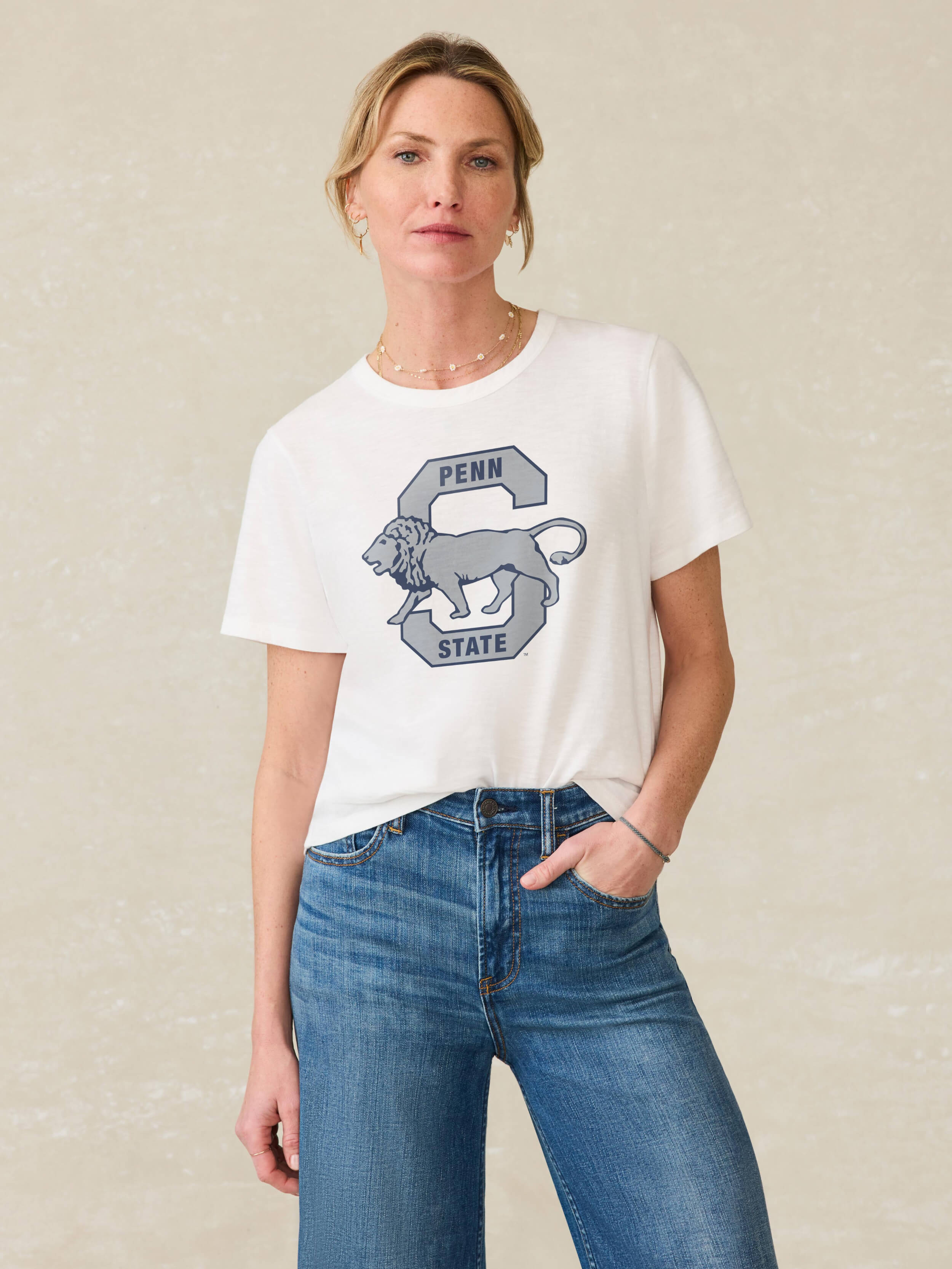 Woman wearing a white t-shirt with a Penn State graphic on the center front and blue jeans against a beige background