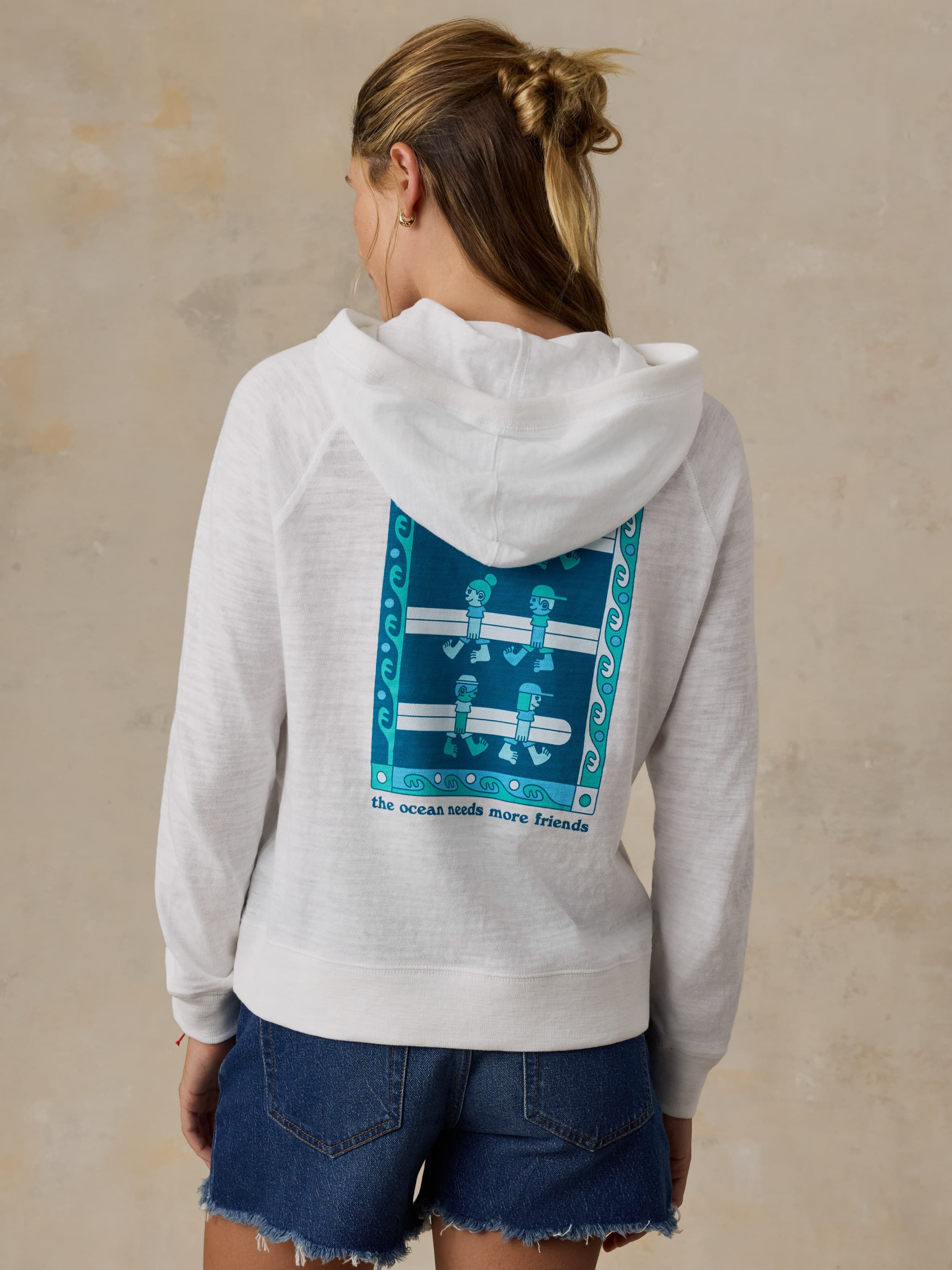 Woman wearing a white Faherty x Surfrider Foundation hoodie against a beige background