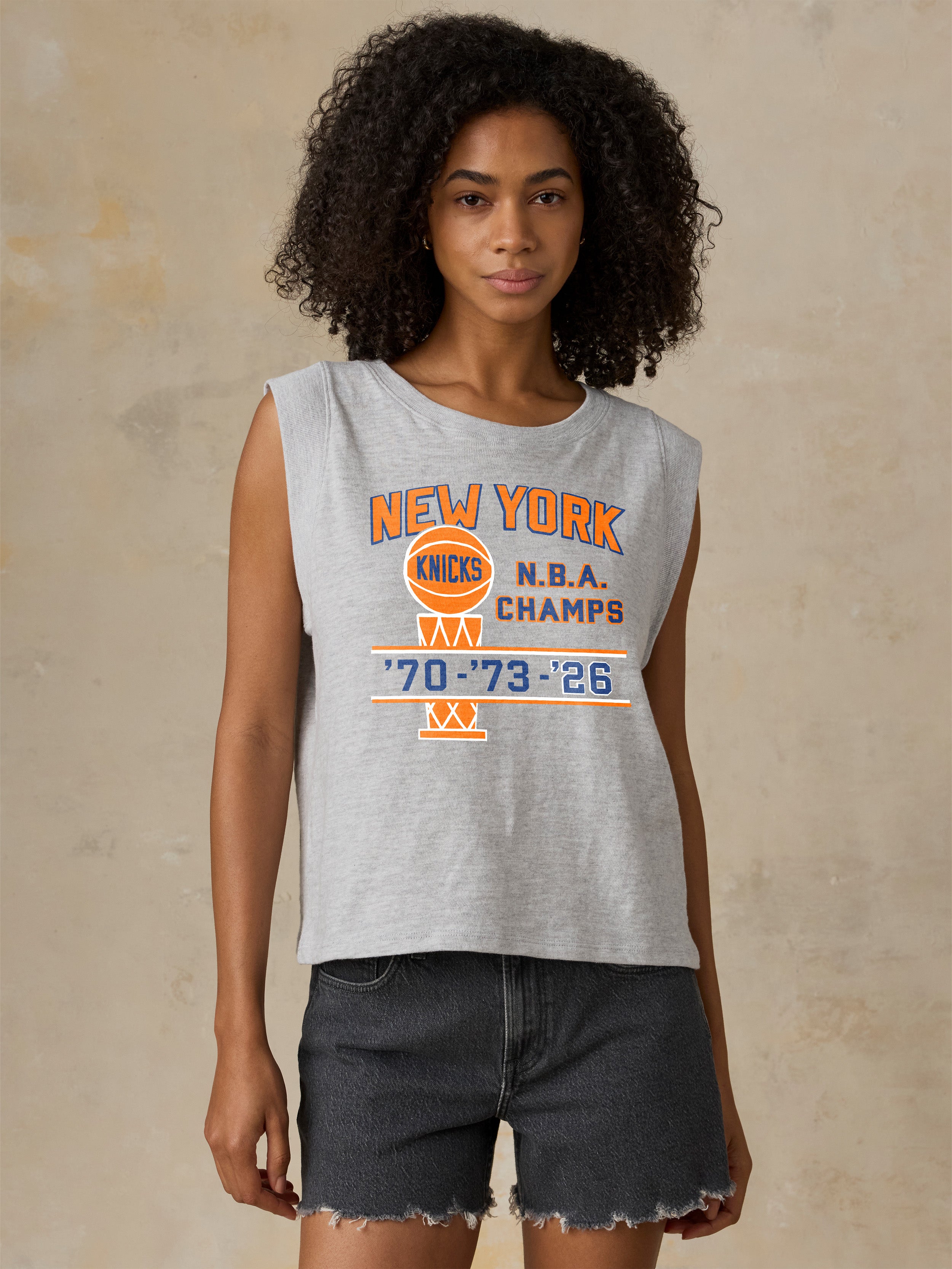 Woman wearing a gray sleeveless shirt with NY Knicks NBA Champs graphic on the center chest against a beige background