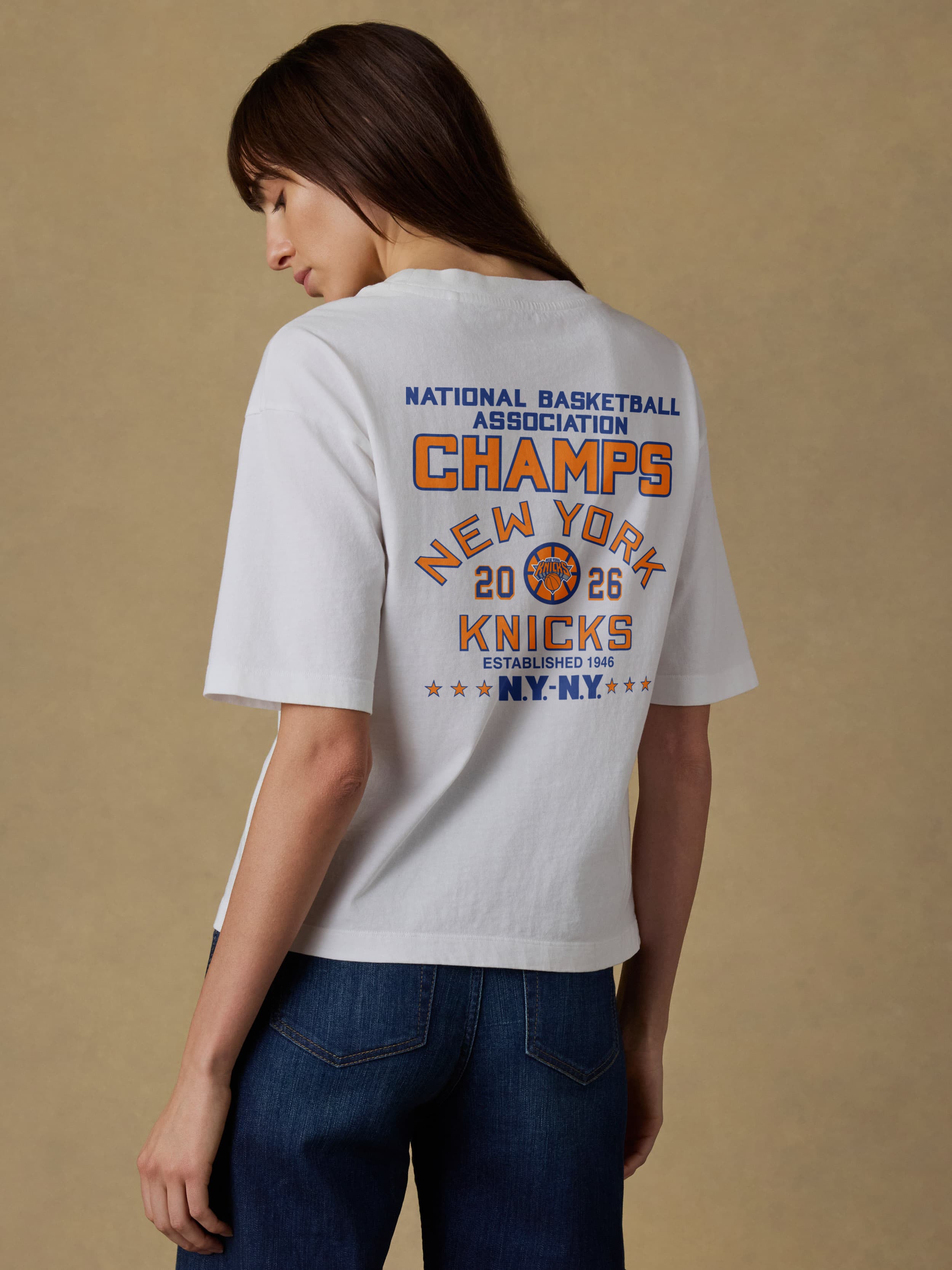 Woman wearing a white t-shirt with New York Knicks championship graphic on the back against a beige background