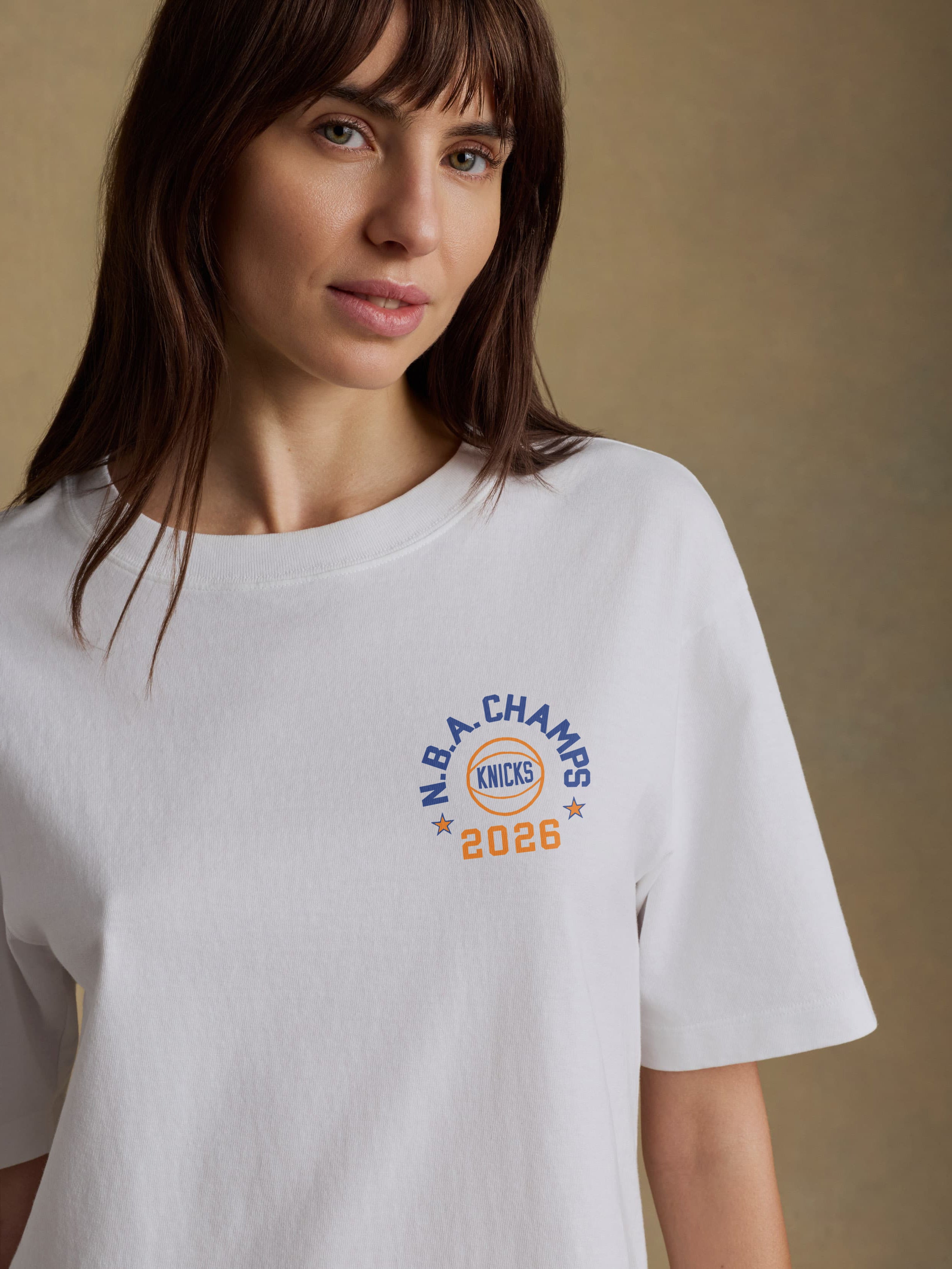 Woman wearing a white t-shirt with 'NY Knicks NBA Champs 2026' graphic on the left chest against on a beige background