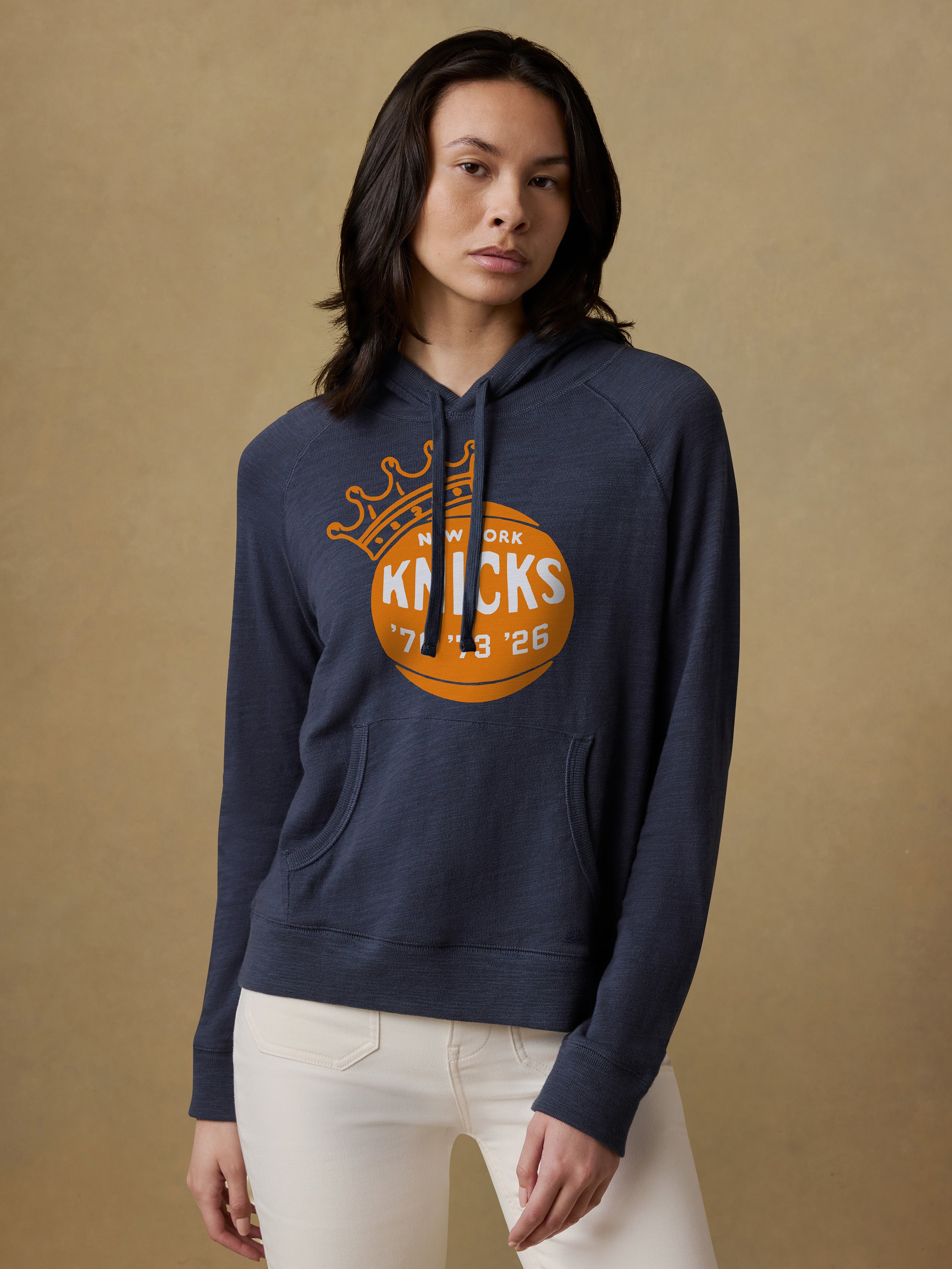 Woman wearing a navy blue hoodie with Knicks graphic on the center chest against a beige background