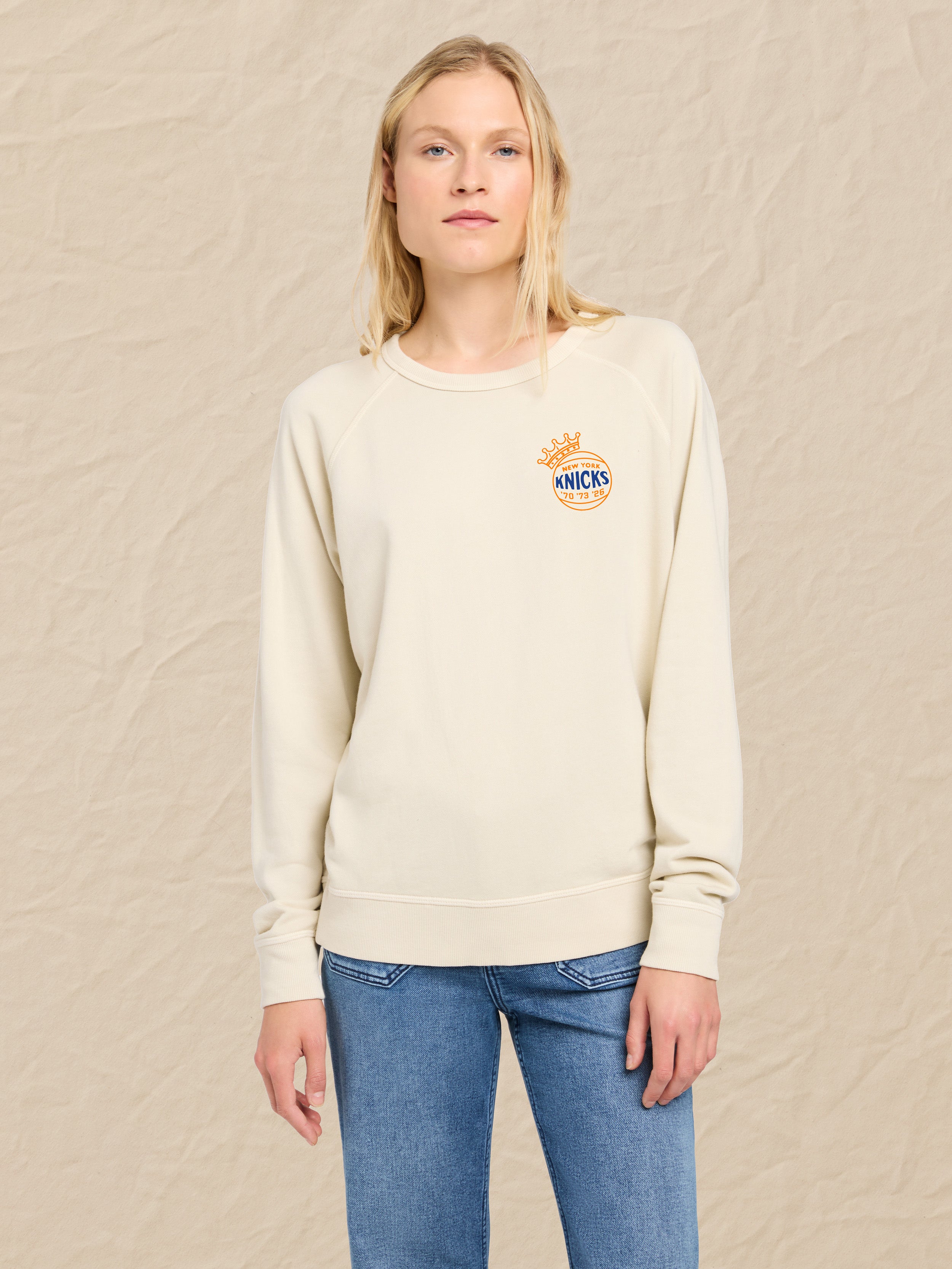 Woman wearing a cream sweatshirt with a NY Knicks graphic on the left chest against a beige background