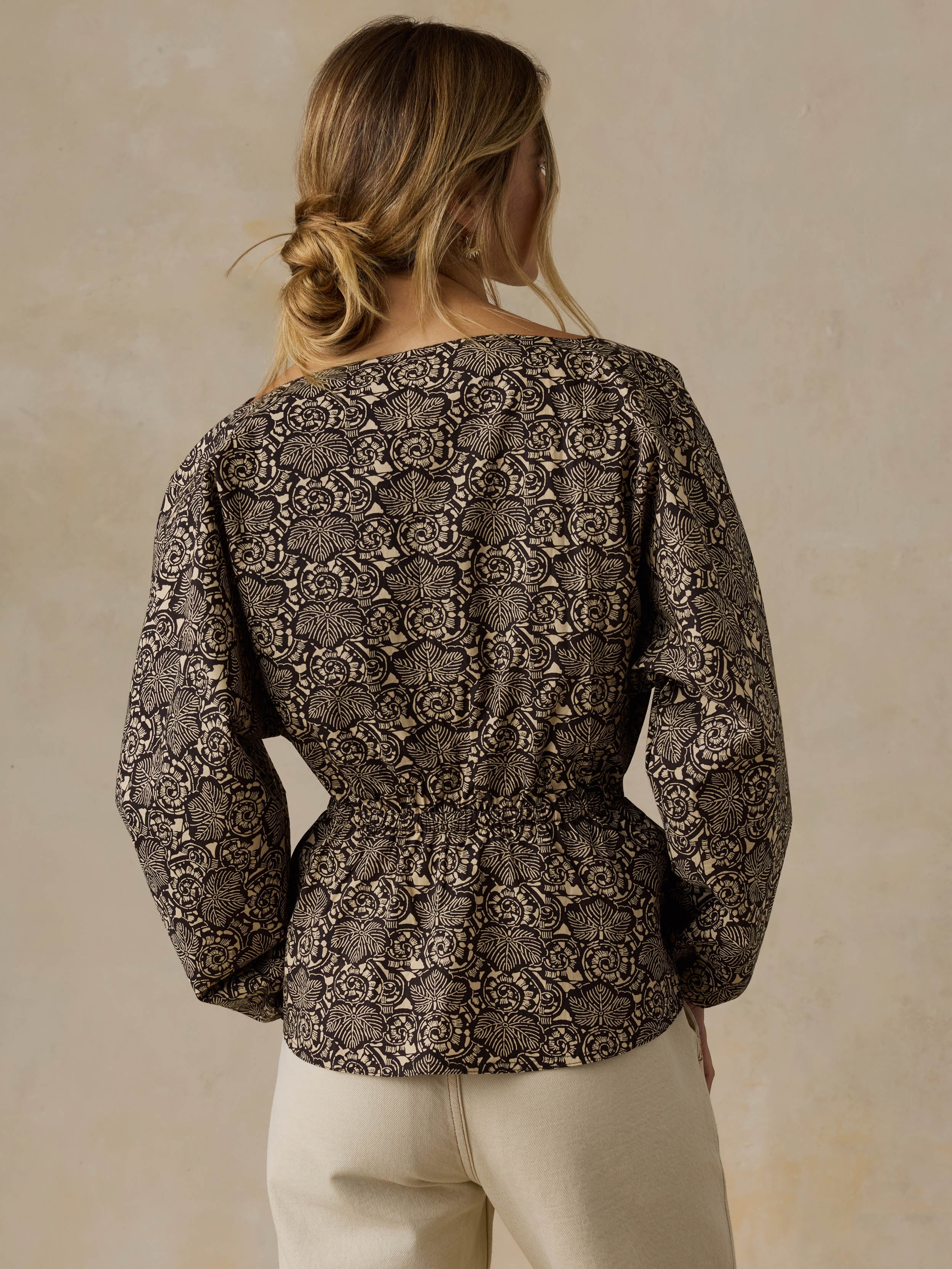 Woman wearing a black patterned blouse and white jeans against a beige background