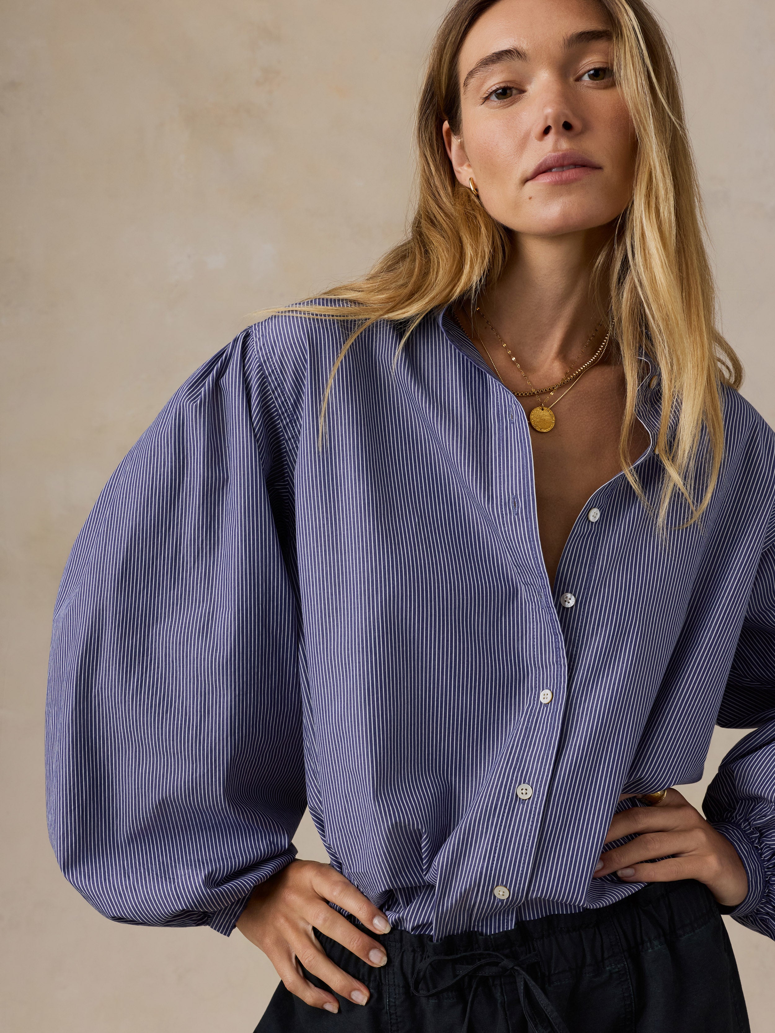 A woman wearing a blue and white vertically striped button-up blouse with dramatic balloon sleeves, paired with dark drawstring trousers, posed against a textured beige wall.