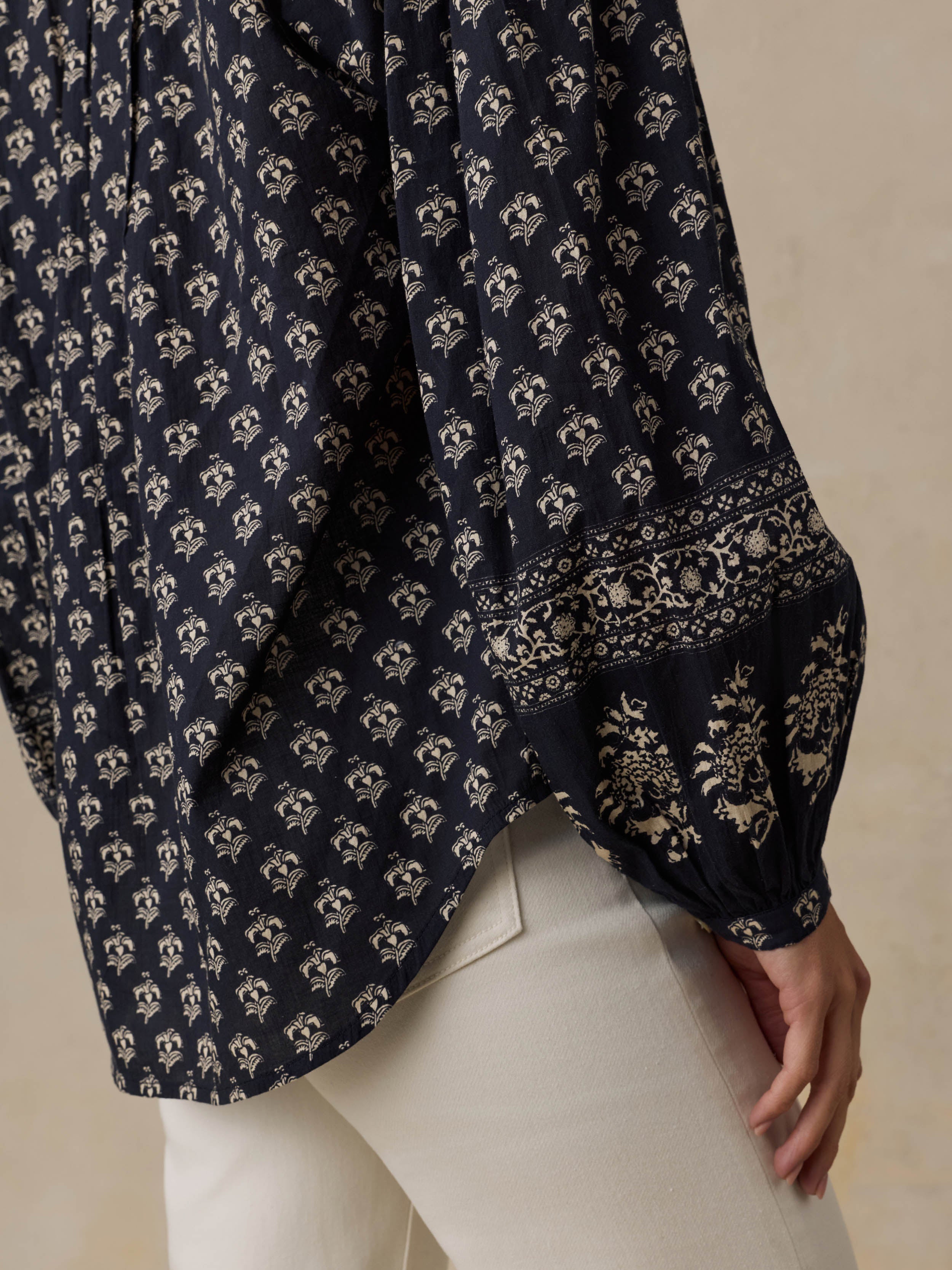 Woman wearing a dark patterned blouse and white pants against a beige background