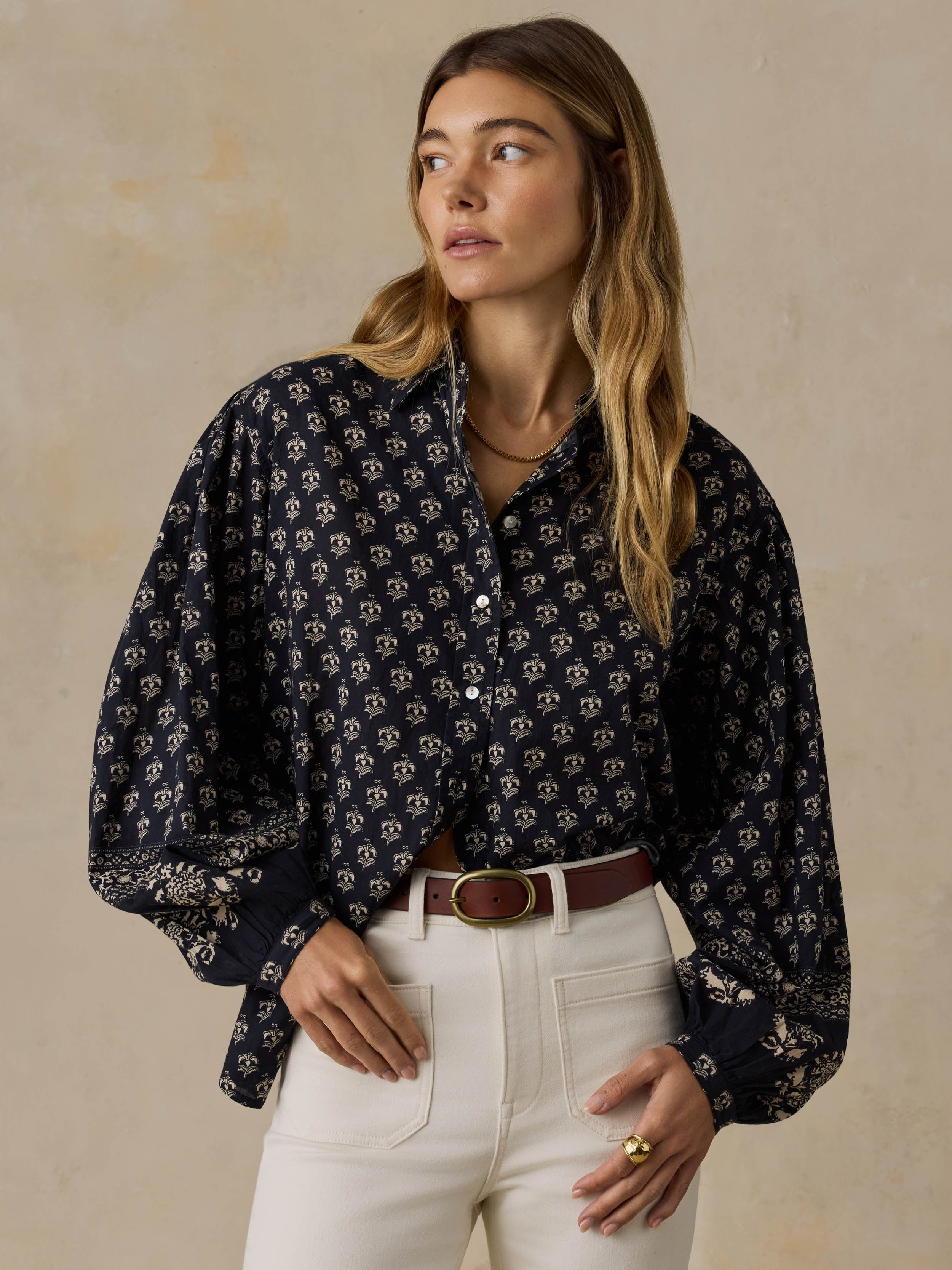 Woman wearing a dark patterned blouse and white pants against a beige background