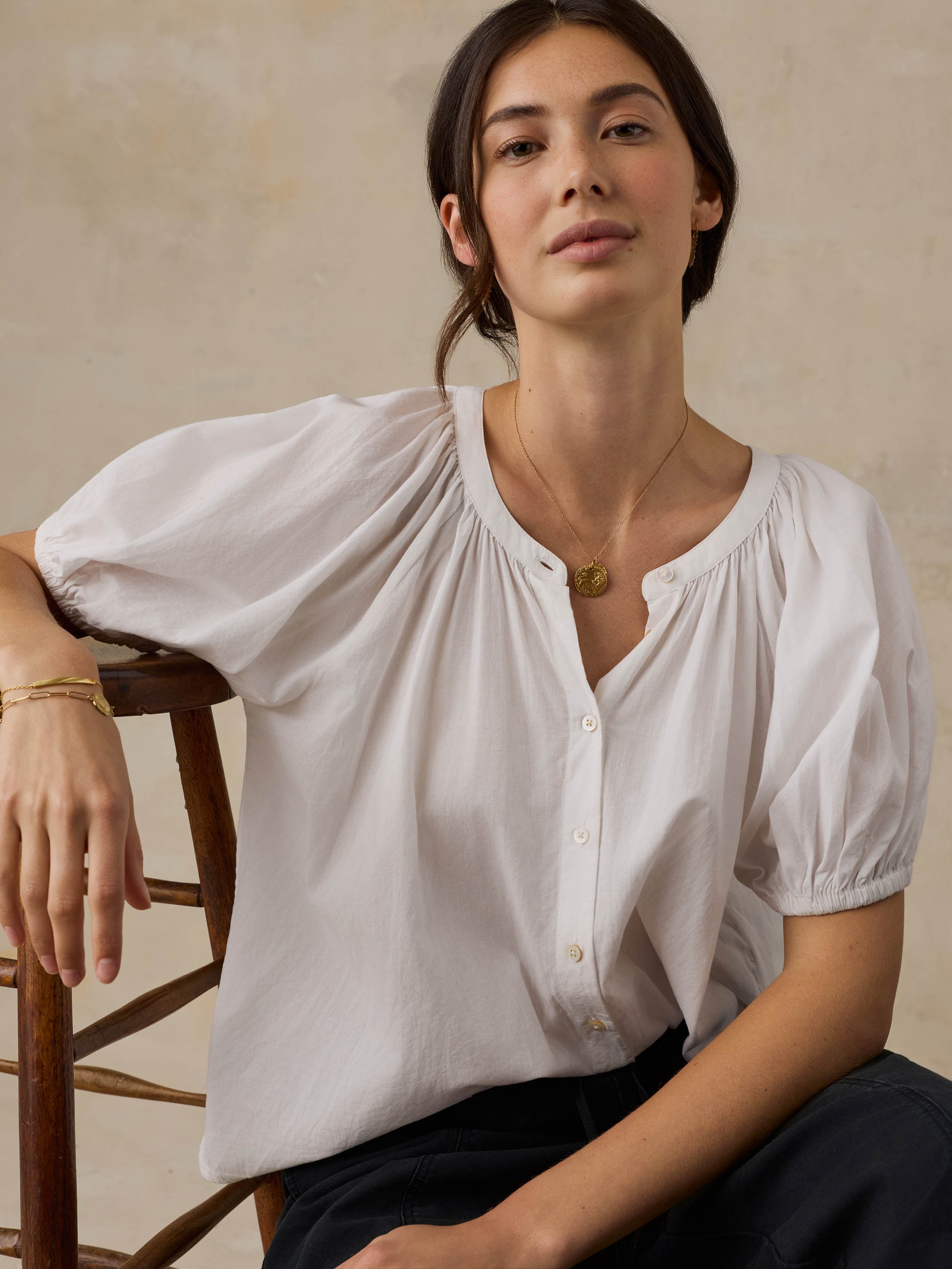 Woman sitting on a wooden chair wearing a white blouse with puff sleeves against a beige background