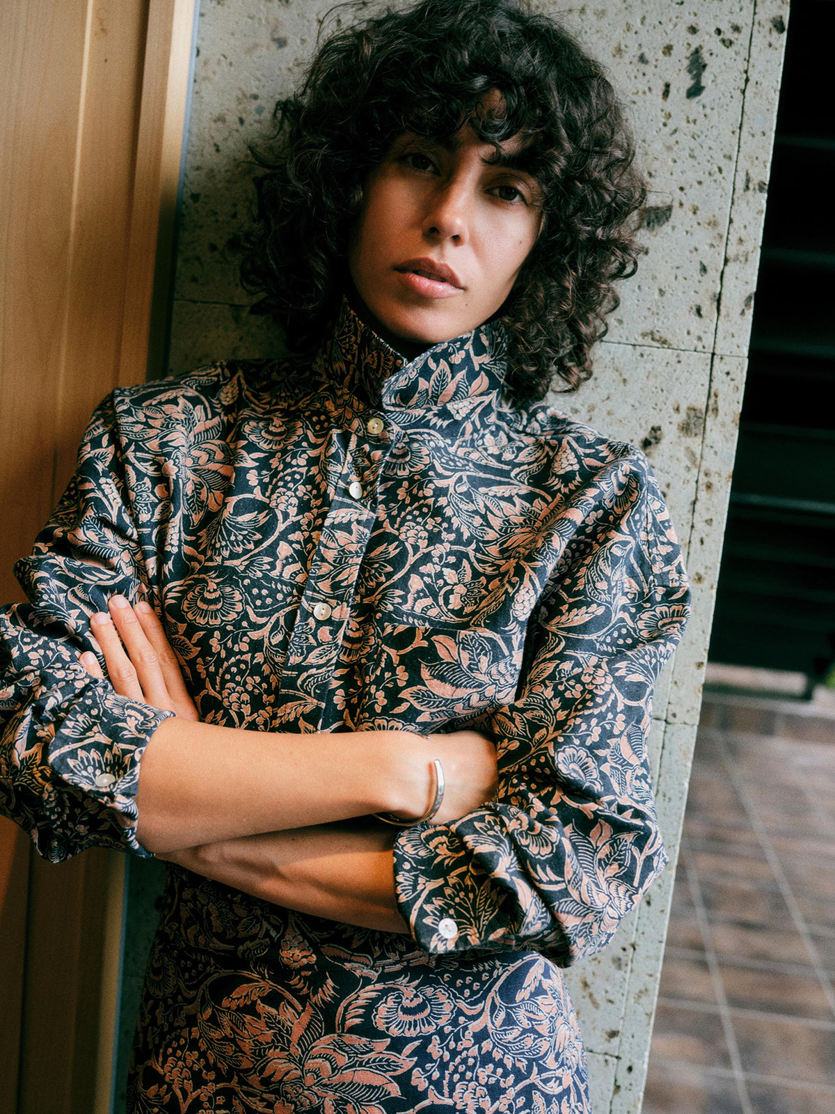A woman with curly dark hair wearing a high-neck, long-sleeve button-up shirt with a dark navy and beige botanical pattern, leaning against a textured concrete pillar., image 1 of 7