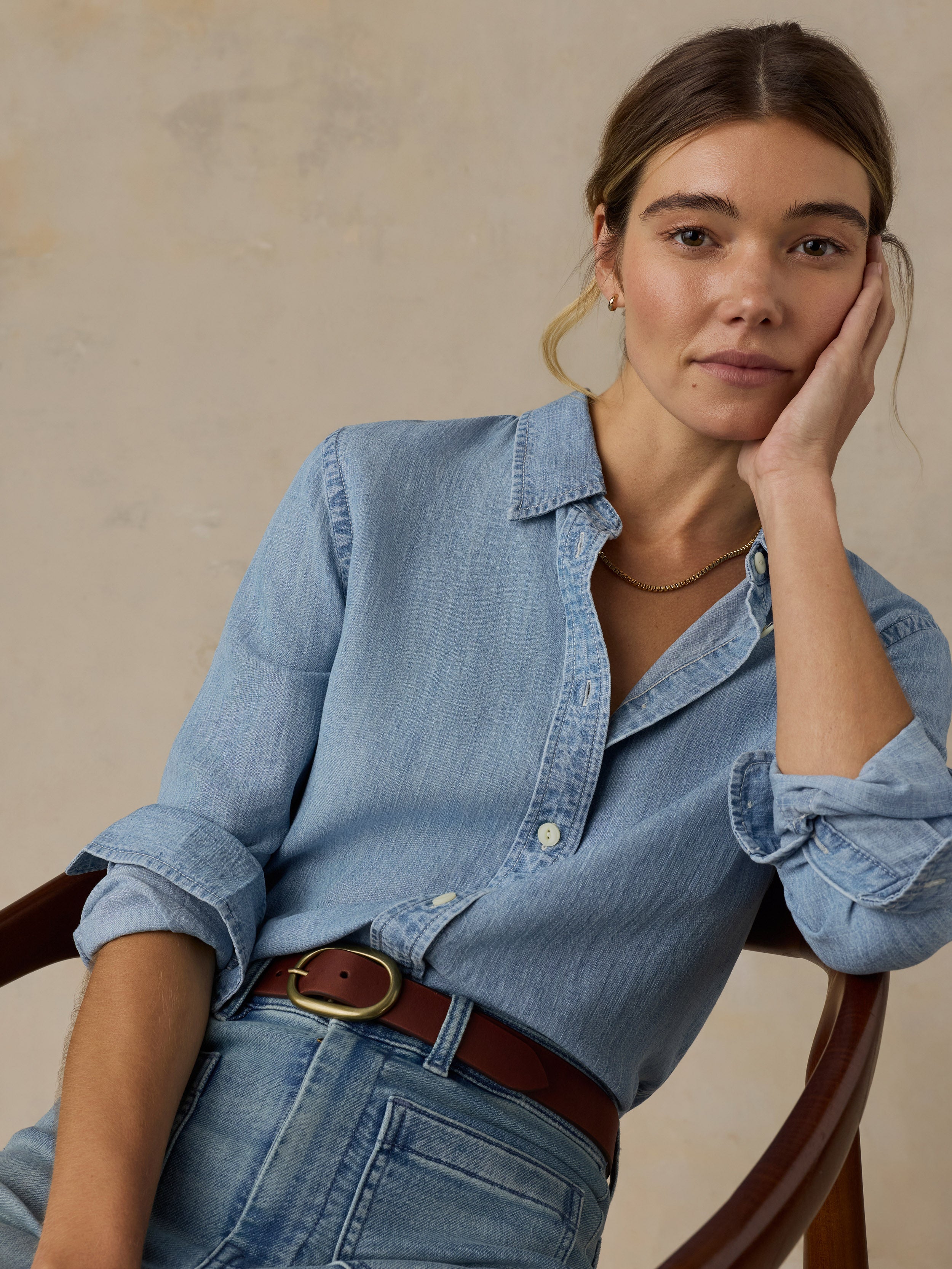 Woman sitting on a wooden chair wearing a chambray button down shirt with blue jeans against a beige background
