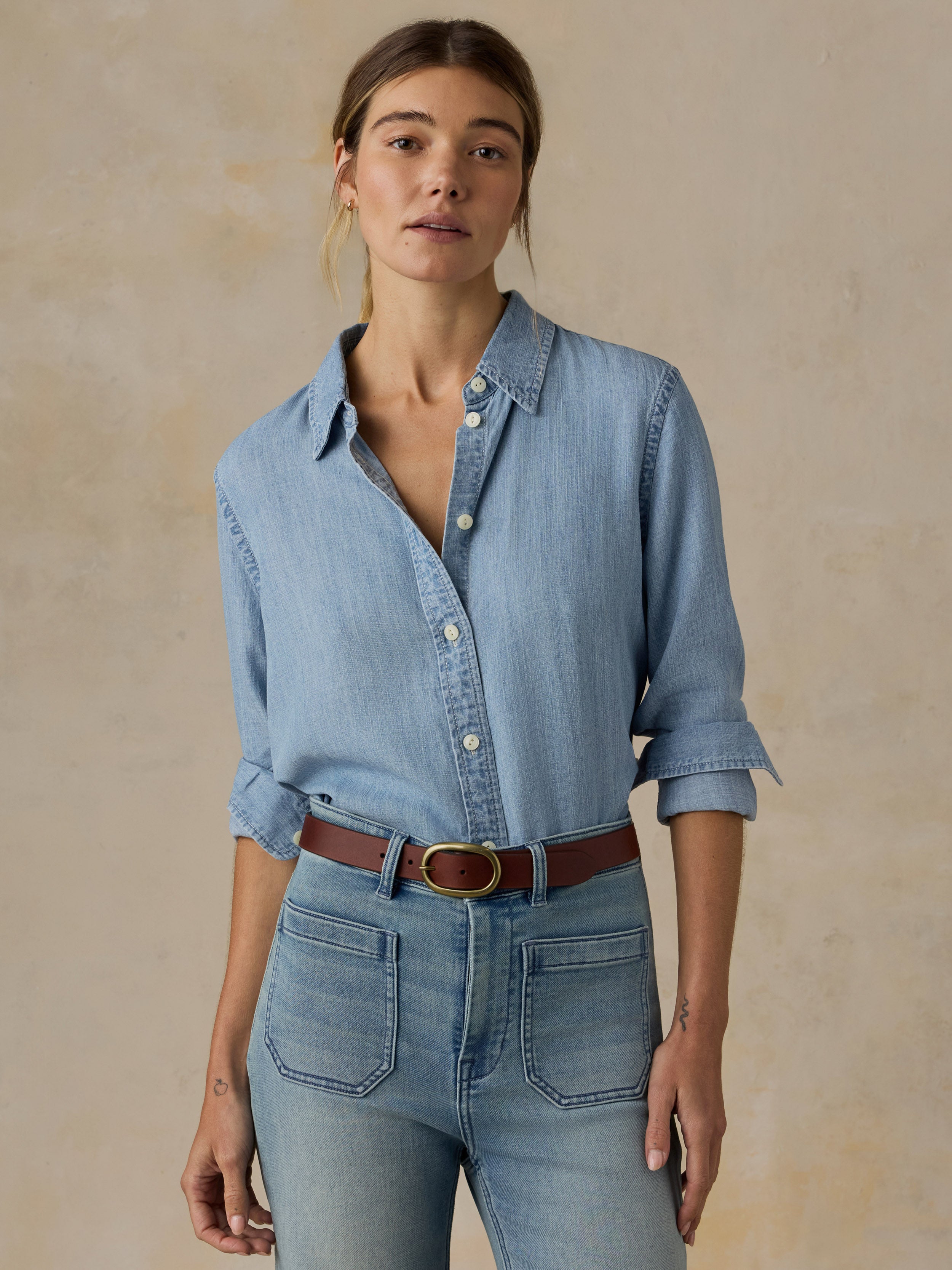 Woman wearing a chambray button down shirt with blue jeans against a beige background