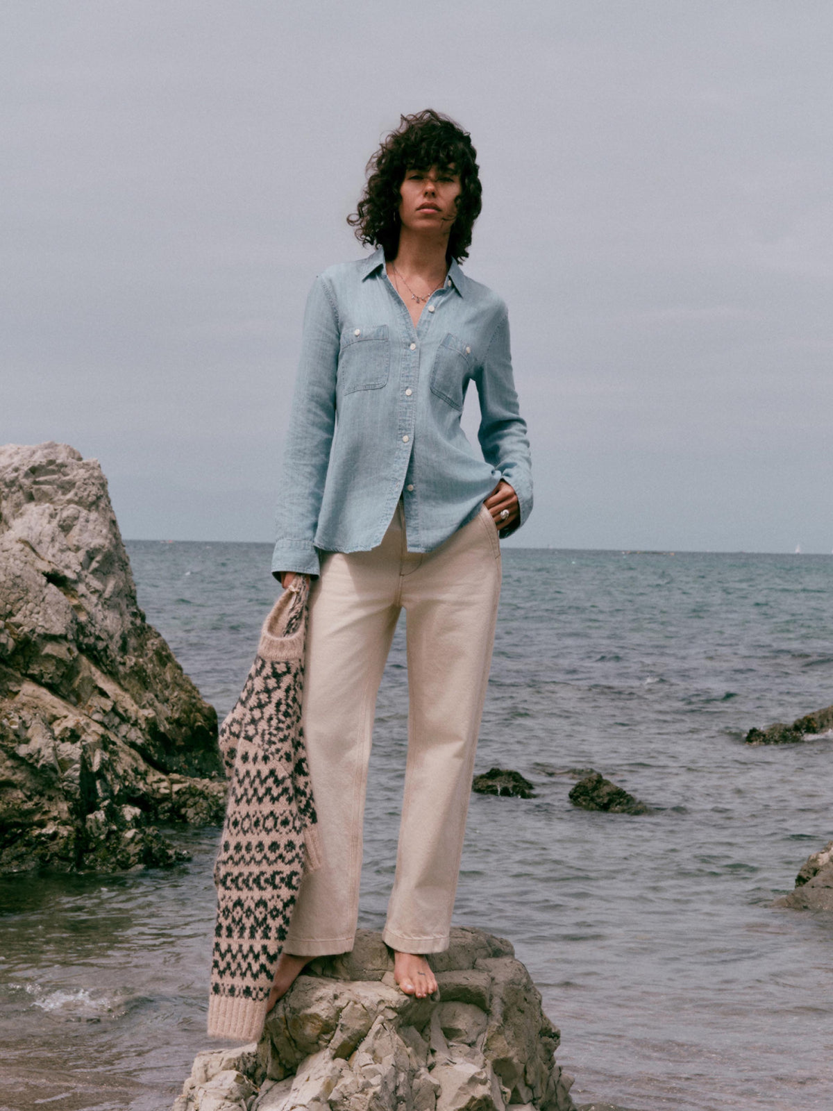 A woman stands barefoot on a rocky shoreline wearing a light blue button-up shirt, cream wide-leg pants, and holding a patterned knit cardigan against an overcast sea background., image 1 of 9