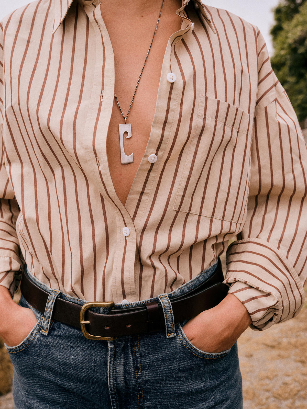 A woman wearing a beige button-up shirt with thin brown vertical stripes, tucked into blue jeans with a dark brown belt. The shirt is unbuttoned at the chest, showing a silver pendant necklace, and the background is a soft-focus outdoor setting., image 1 of 6