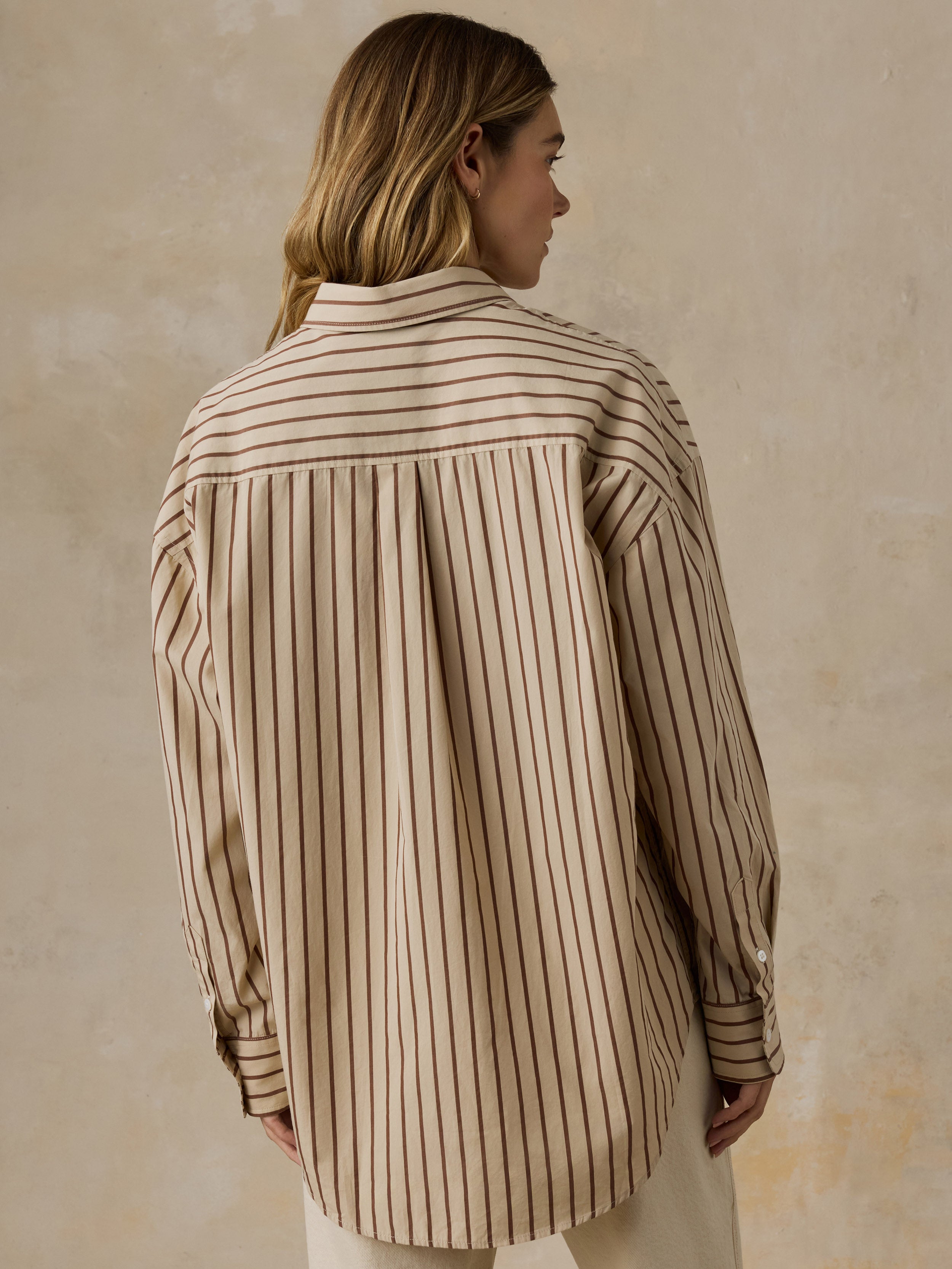 Woman wearing a beige and brown striped button down shirt and white jeans against a beige background