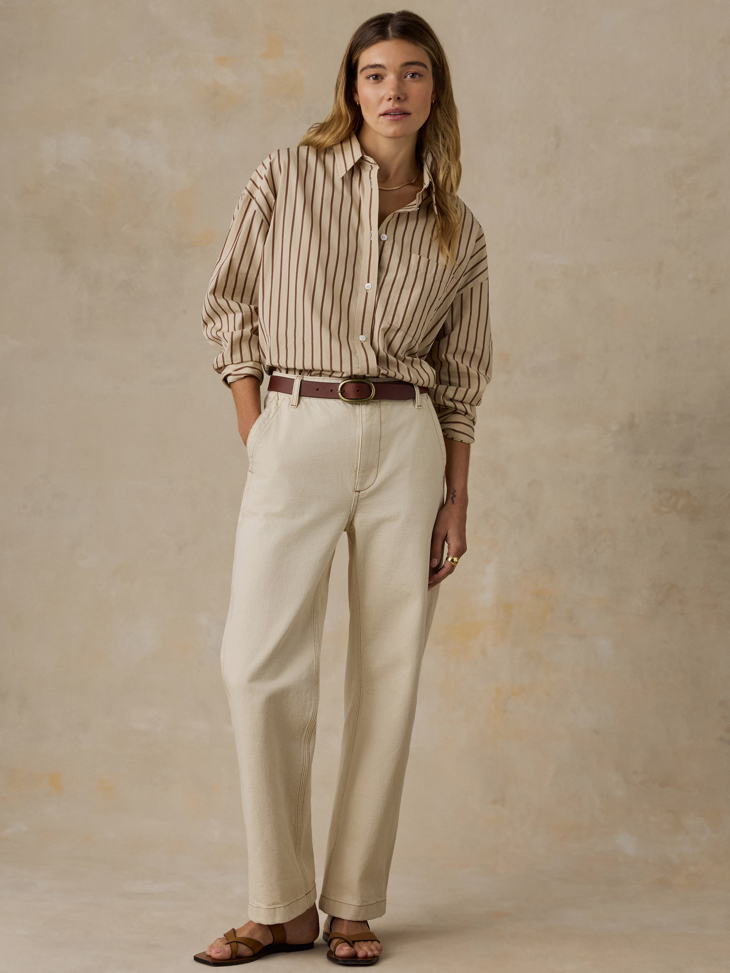Woman wearing a beige and brown striped button down shirt and white jeans against a beige background