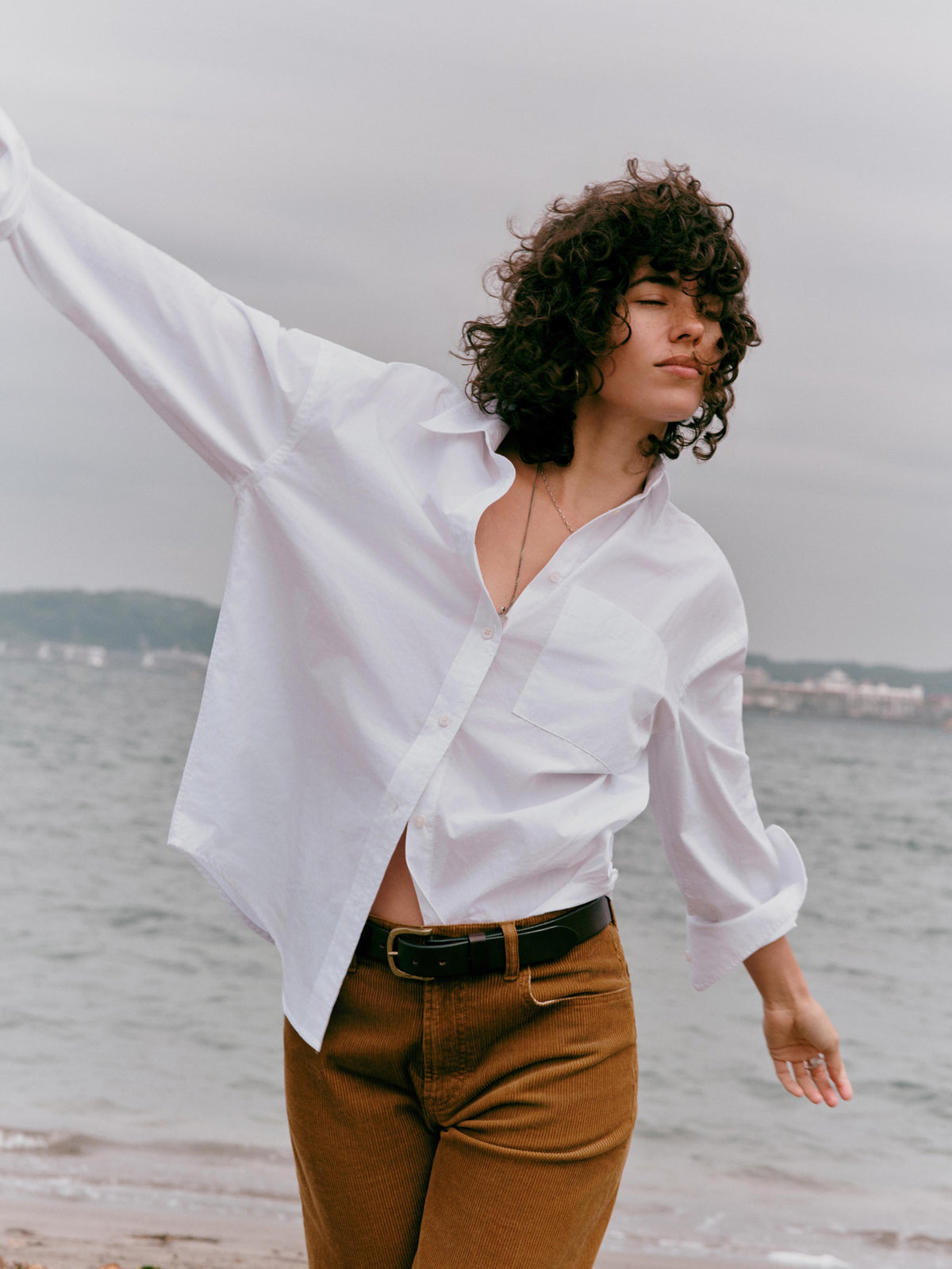 A woman with curly dark hair stands on a beach wearing an oversized white button-down shirt and brown pants, with her arm raised against a cloudy sky and water in the background., image 1 of 7