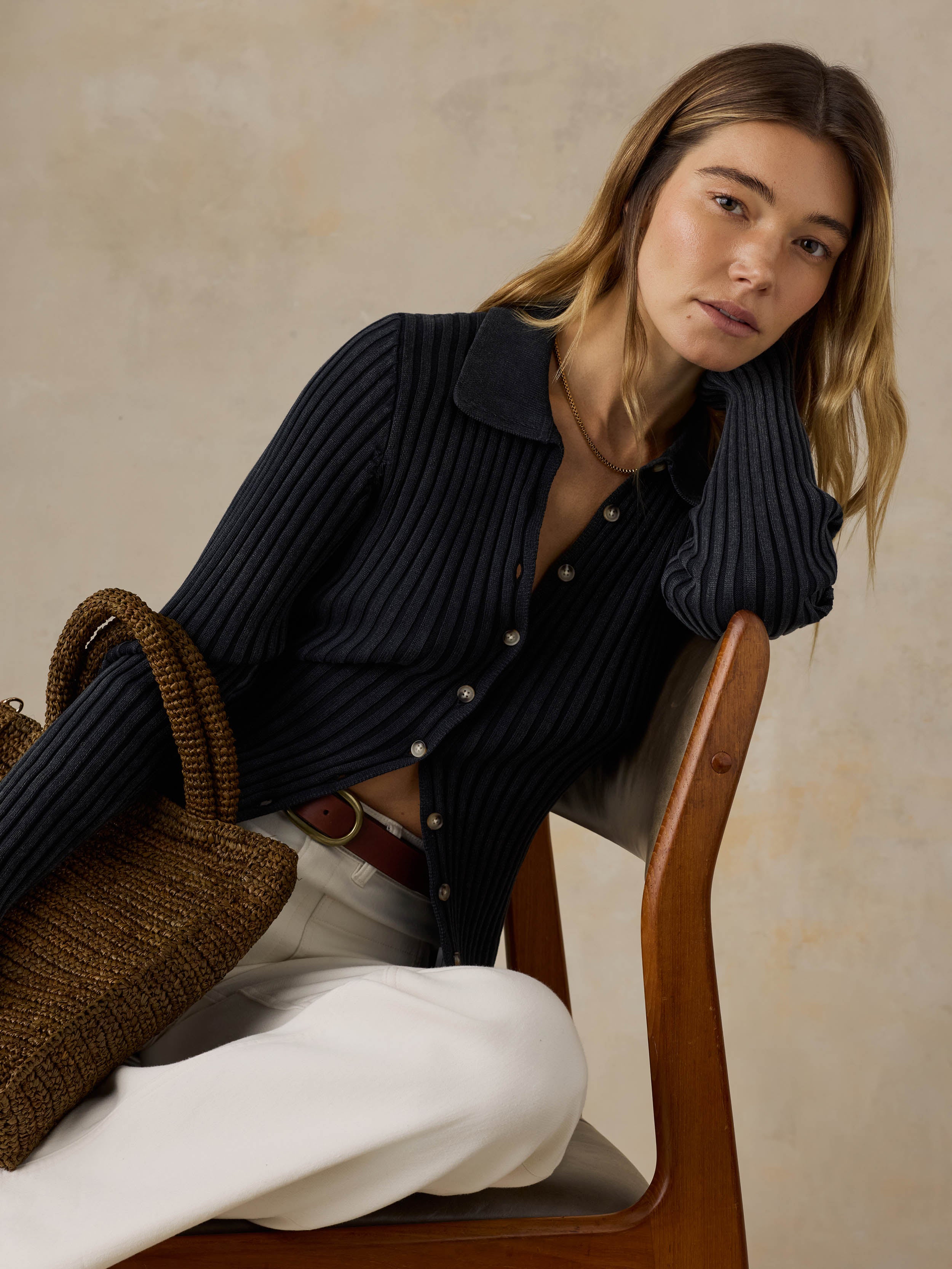 Woman sitting on a wooden chair wearing a black ribbed cardigan and white pants against a beige background